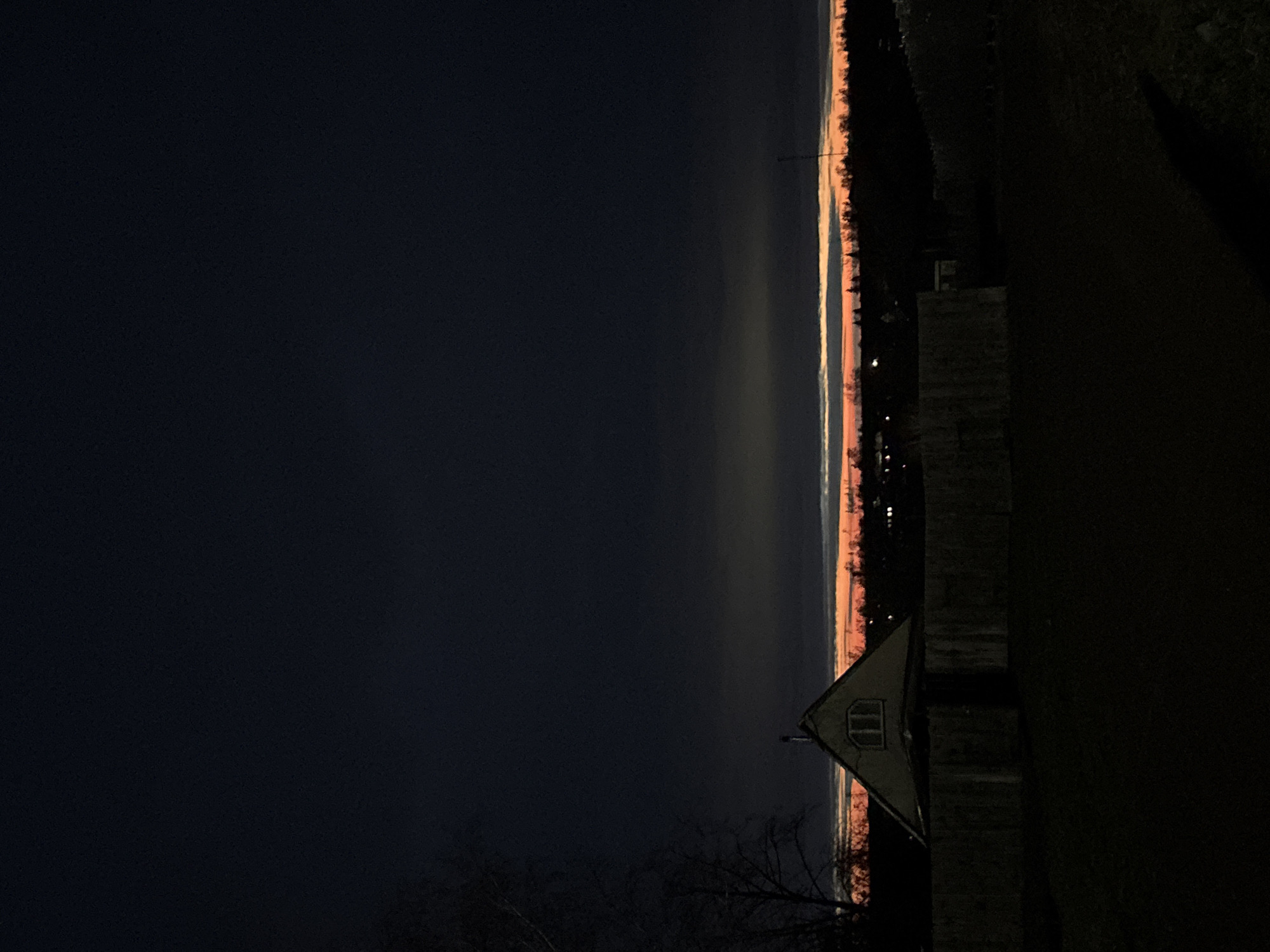 The image displays a twilight or early morning landscape, likely capturing either a sunrise or sunset. The upper two-thirds of the frame are dominated by a dark, deep blue-black sky. A striking horizontal band of bright orange, pink, and light blue stretches across the horizon, signifying the position of the sun just below the visible scene. Below this vibrant horizon, a dark, undulating line of distant land or trees is silhouetted.
In the lower right foreground, a small, dark-silhouetted building, possibly a shed or a house, is visible. It features a distinct triangular roof and a single, small rectangular window with visible mullions. Adjacent to this structure, on the far right, a solid, dark vertical mass, perhaps a wall or fence, is present. In the lower left corner, sparse, bare tree branches reach upwards into the dark sky.
No people are visible, and no specific activity is taking place. The time of day is clearly either dawn or dusk, emphasized by the dramatic colors on the horizon and the overall low light. The scene evokes a sense of tranquility and solitude. There is no visible text. The setting appears to be a rural or semi-rural area, consistent with the provided location context of Фоки, Russia.