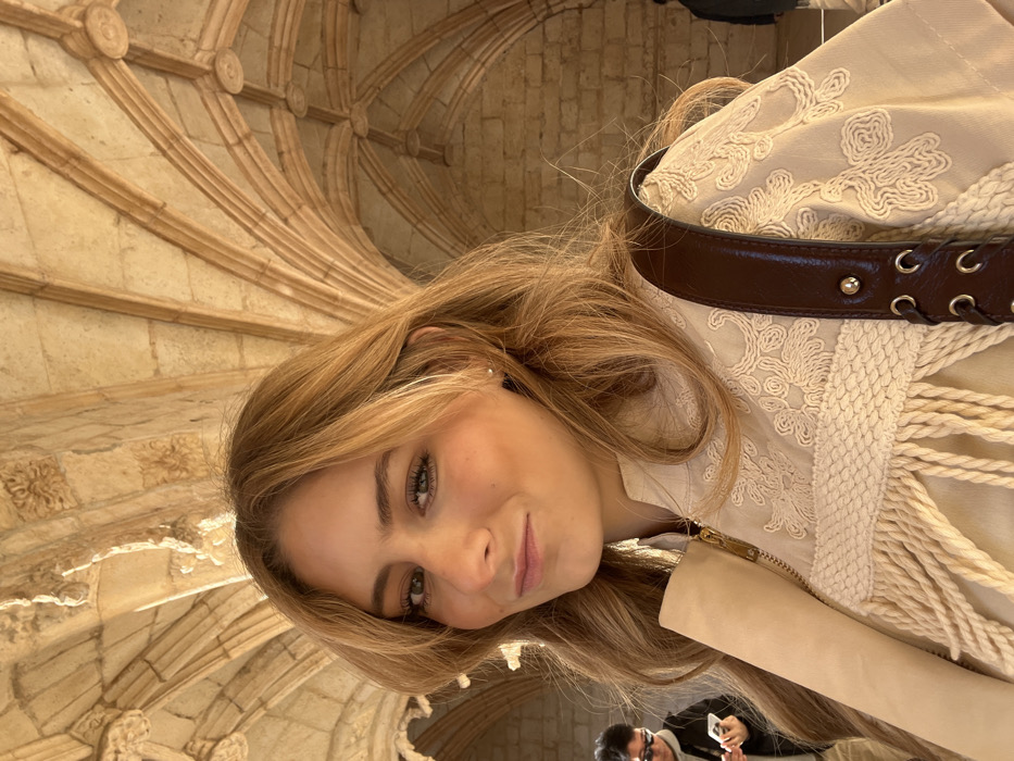 The image depicts a young Caucasian female with long, wavy blonde hair and green eyes, looking directly at the camera with a subtle smile. She occupies the central foreground, wearing a light beige jacket adorned with intricate white embroidery, a woven horizontal band, and cream-colored fringe details. A gold zipper is visible on her jacket. A dark brown leather shoulder strap of a bag rests on her right shoulder. Her left ear displays a small, spherical earring.
In the blurred background, to the left, a person with dark hair and sunglasses is partially visible, looking down at a white smartphone. A second, indistinct figure is present in the far right background.
The architectural setting features elaborate light-colored stone, showcasing complex rib vaulting and decorative bosses characteristic of the Manueline style. This specific architectural detail firmly places the location within the cloisters of the Jerónimos Monastery (Mosteiro dos Jerónimos) in Belém, Lisbon, Portugal. The lighting is bright and natural, indicating an open-air or well-lit semi-enclosed space.