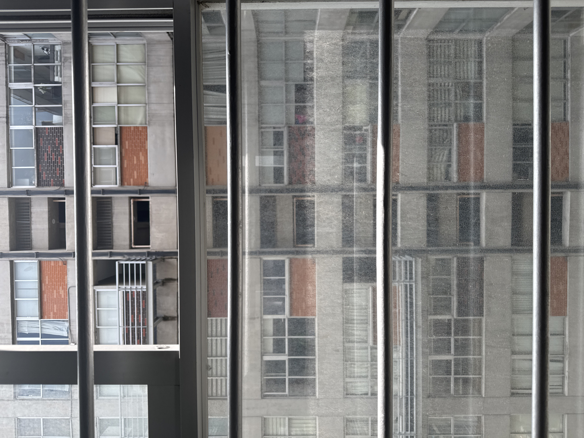 The image shows a close-up view of the exterior of a multi-story building, as seen through a window with horizontal bars. The building appears to be made of concrete and brick, with numerous windows and balconies. The windows are of various sizes and styles, some with curtains or blinds visible. There are horizontal metal bars in the foreground, obscuring parts of the view. The building's architecture suggests an urban setting, possibly an apartment complex or office building. The lighting indicates it is daytime.