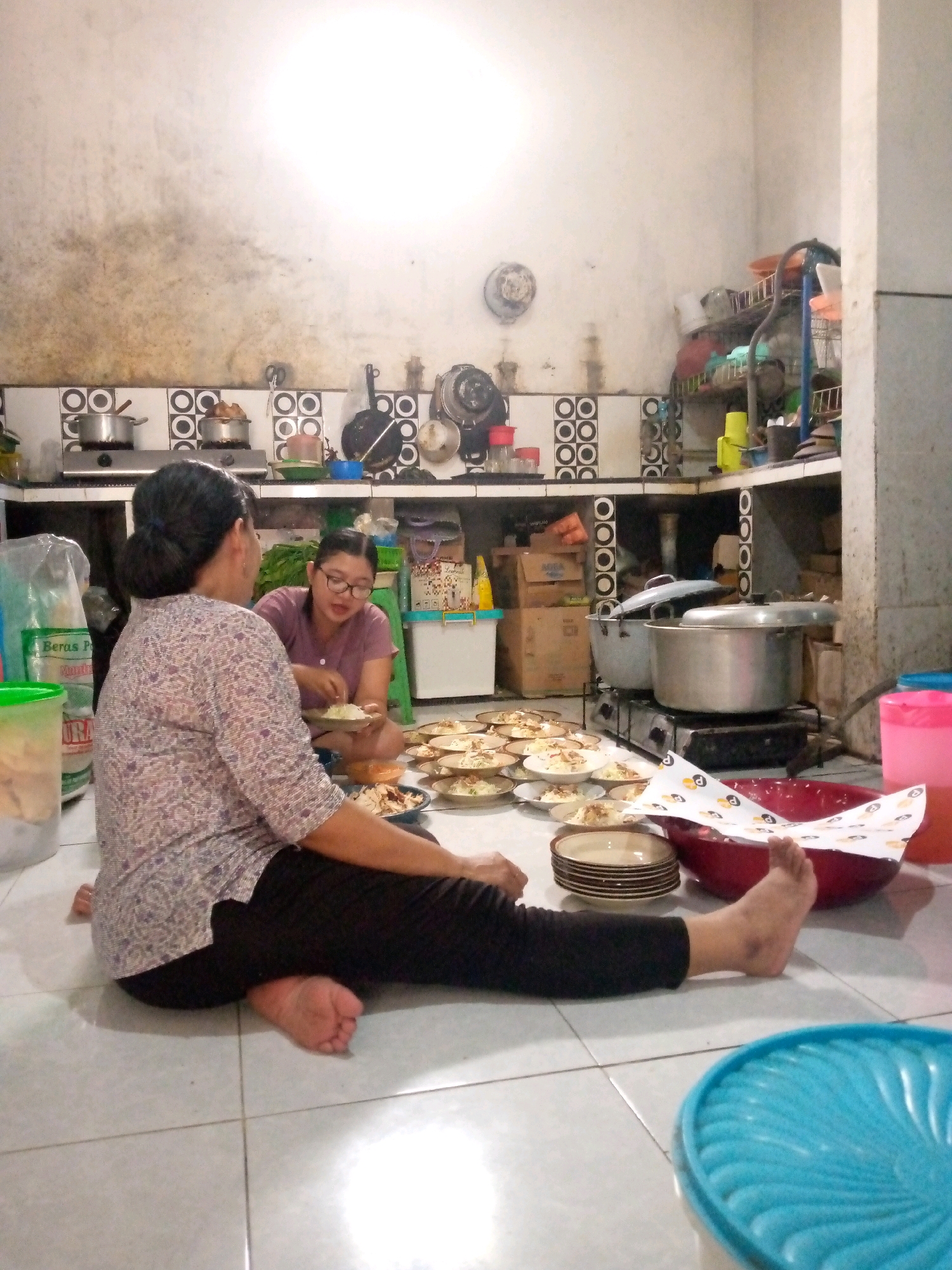 In a kitchen located in Sedati, Indonesia, two women are preparing food. One woman, seated on the tiled floor with her back to the camera, wears a patterned blouse and black pants. The other woman, wearing a purple shirt and glasses, sits opposite her, engaged in serving food onto plates. The floor is covered with numerous plates of food, likely prepared for an event. The kitchen environment appears utilitarian, with cooking equipment, shelves stocked with items, and visible plumbing. The lighting is somewhat dim, suggesting it might be evening.