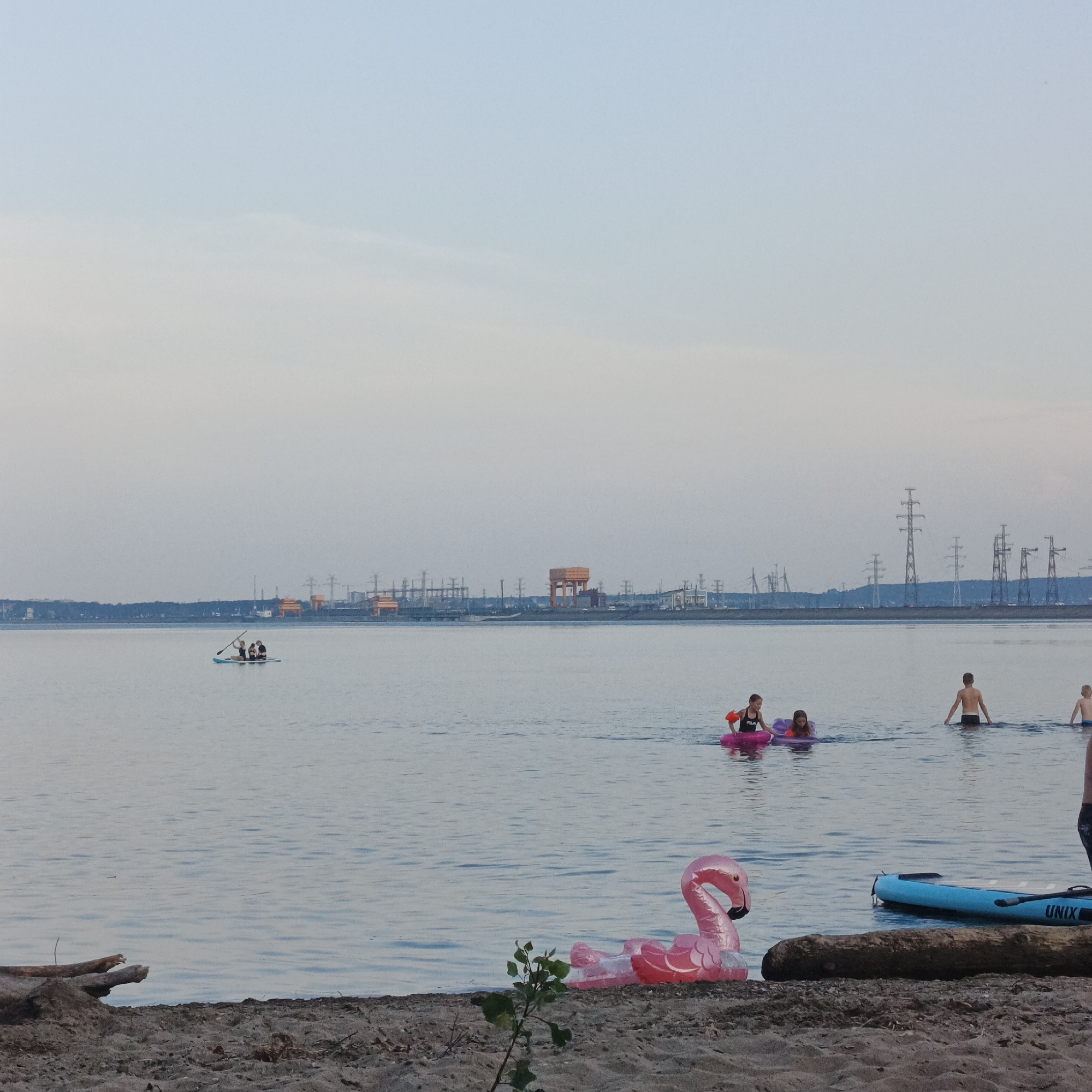 In Noviy, Russia, a serene body of water serves as a recreational area for a diverse group of individuals. The scene is dominated by a calm expanse of water, meeting a sandy shoreline in the foreground. On the beach, a pink inflatable flamingo float rests near a large piece of driftwood, with a small plant growing nearby.
Further out in the water, several people are engaged in various activities. A paddleboard, blue with the "UNIX" logo visible, lies near the shore. A group of people are seen on a paddleboard in the distance, paddling with oars. Closer to the shore, children play in the water, with two girls on inflatable rings. One girl wears a black and red swim top with orange armbands, and the other is on a purple float. A young boy stands in the water further out, facing away from the camera. Another child is partially visible in the distance.
The backdrop features a sprawling industrial landscape, characterized by numerous electrical transmission towers and large industrial structures, some with orange accents. The sky above is a soft gradient of pale blue and grey, suggesting either early morning or late evening light. The overall impression is one of peaceful coexistence between natural recreation and the industrial environment.