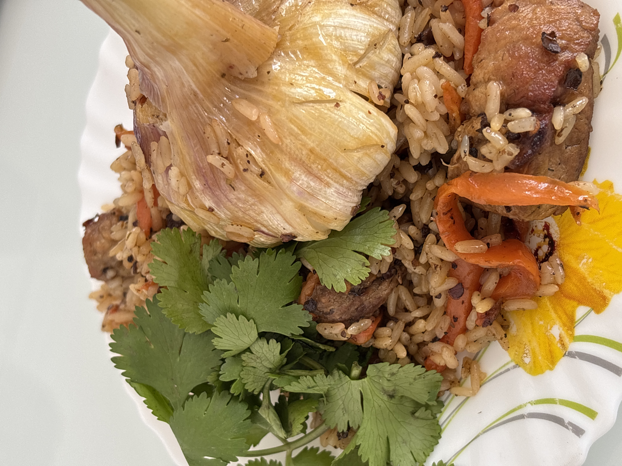 A plate of pilaf, garnished with cilantro and a roasted head of garlic, rests on a white surface. The pilaf contains rice, carrots, and meat. The plate features a floral design. The scene is indoors, likely in a private home. The location in Prikamskii, Russia is assumed, as it was provided in the instructions. There are no people visible in the image.