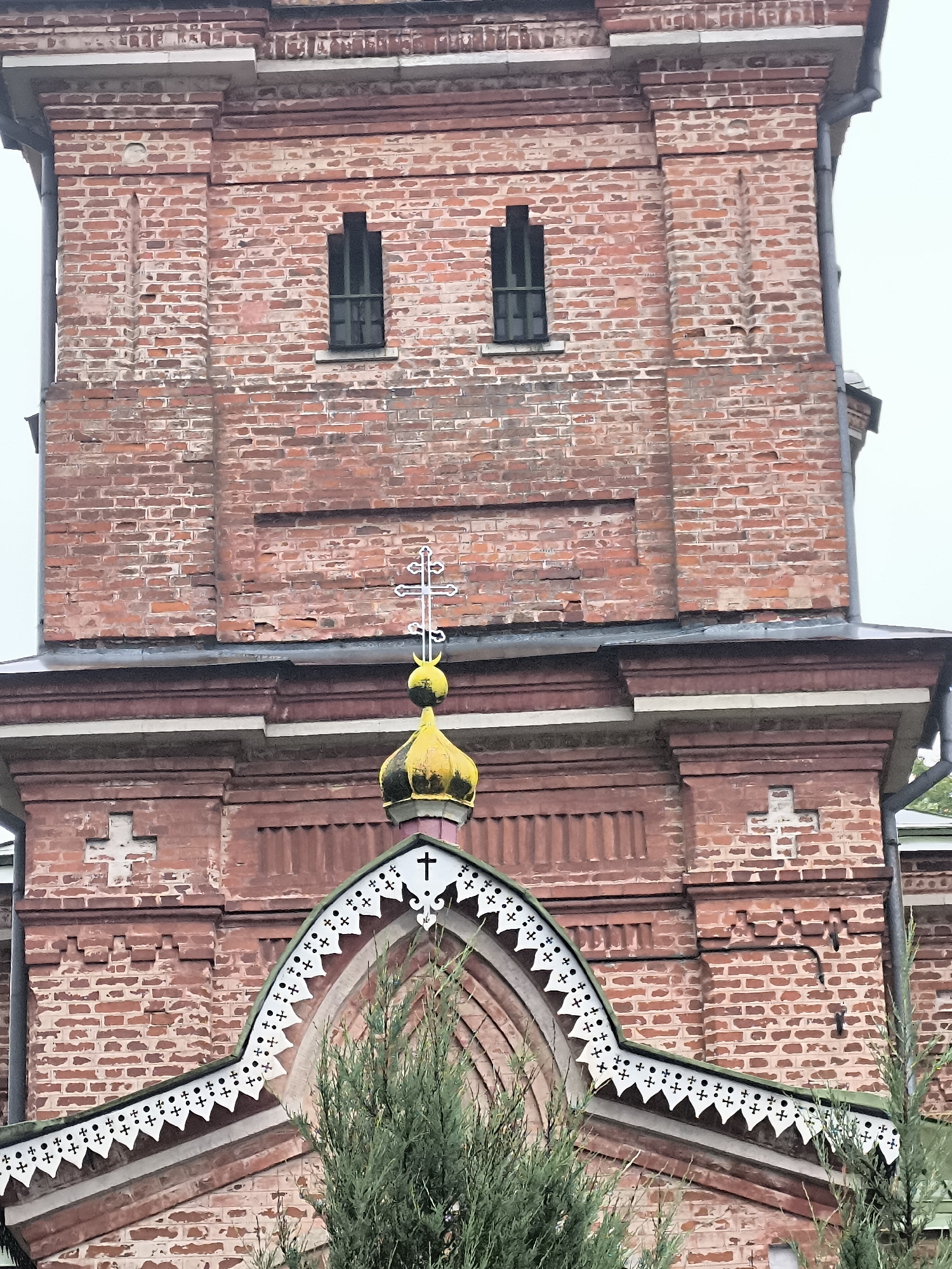 The image shows a close-up of a red brick building, likely a church, with ornate architectural details. The building features two narrow, dark-colored windows positioned vertically on the upper section. Below the windows, a gold-colored dome with a silver cross on top is visible, situated above a pointed archway with white decorative trim. The brickwork exhibits signs of age and weathering, with some bricks appearing chipped or discolored. The surrounding environment is not clearly visible, but a hint of green foliage suggests it might be an outdoor setting. This building is identified as part of the Holy Trinity Orthodox Church in Kuldīga, Latvia.