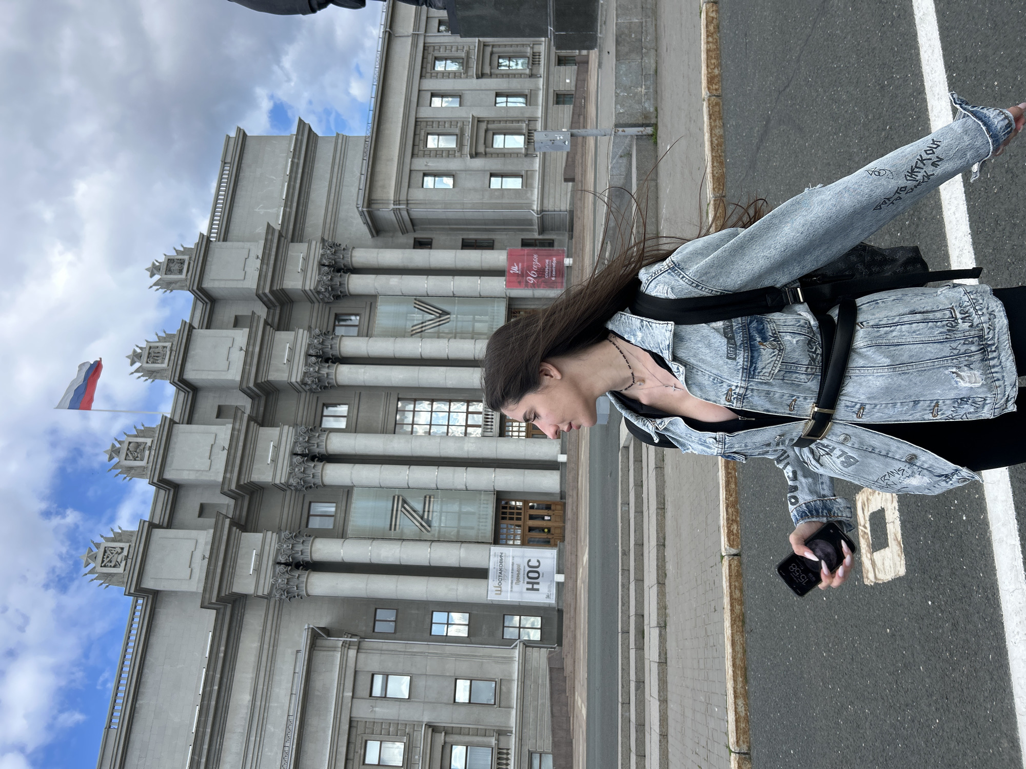 This media file shows a young woman standing on a sidewalk in front of a large, imposing stone building with classical architectural elements. The building features grand columns and multiple stories with evenly spaced windows. A Russian flag is visible flying from the top of the building on the left side. The woman is wearing a denim jacket and appears to be holding a smartphone in her hand. She has long, dark hair that is blowing in the wind.
In the background, there is a sign with Cyrillic text. One part of the sign reads "96" followed by some other words and then "создания," which translates to "creation" or "establishment." Another sign below it has the letters "НОС" on it. The building appears to be a government or institutional building, possibly a museum or cultural center, given the signage and architectural style.
The scene is outdoors, and the sky is partially cloudy, suggesting it could be daytime, possibly late afternoon, as indicated by the time displayed on the phone: 15:38. The woman's posture and the slight movement of her hair suggest a gentle breeze. The overall mood is somewhat neutral, with the focus on the juxtaposition of the modern person against the historical architecture. The location is identified as Novokuybyshevsk, Russia.