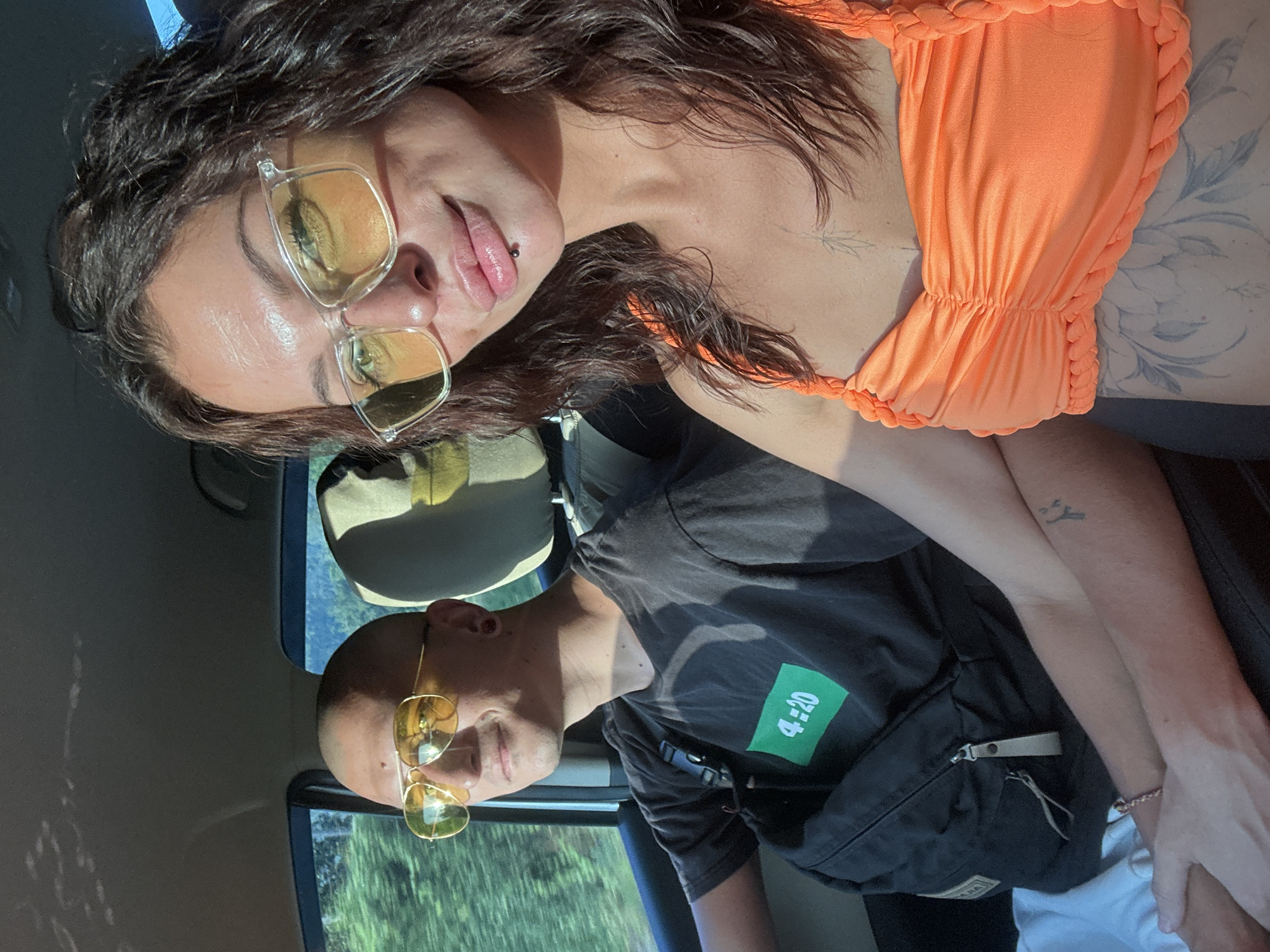 Two individuals, a man and a woman, are seated in the front passenger seats of a vehicle. The man, on the left, is wearing a black t-shirt with a green rectangular graphic displaying "4:20", sunglasses with yellow lenses, and has a shaved head. He is looking forward with a slight smile. A black backpack with a zipped compartment is slung over his shoulder. The woman, on the right, is wearing an orange ruched top and clear-framed sunglasses with yellow lenses. She is looking towards the camera, with her lips slightly parted. She has a lip piercing. Both individuals appear to be in motion, indicated by the blurred greenery visible through the car window, suggesting the vehicle is traveling through a rural or natural setting. The lighting suggests it is daytime, possibly late afternoon due to the warm sunlight. The presence of trees and foliage outside the window indicates a location with lush vegetation. The interior of the car shows car seats and a portion of the dashboard. This scene is likely within the Ko Samui District, Thailand, given the visual cues of tropical foliage and common vehicle types.