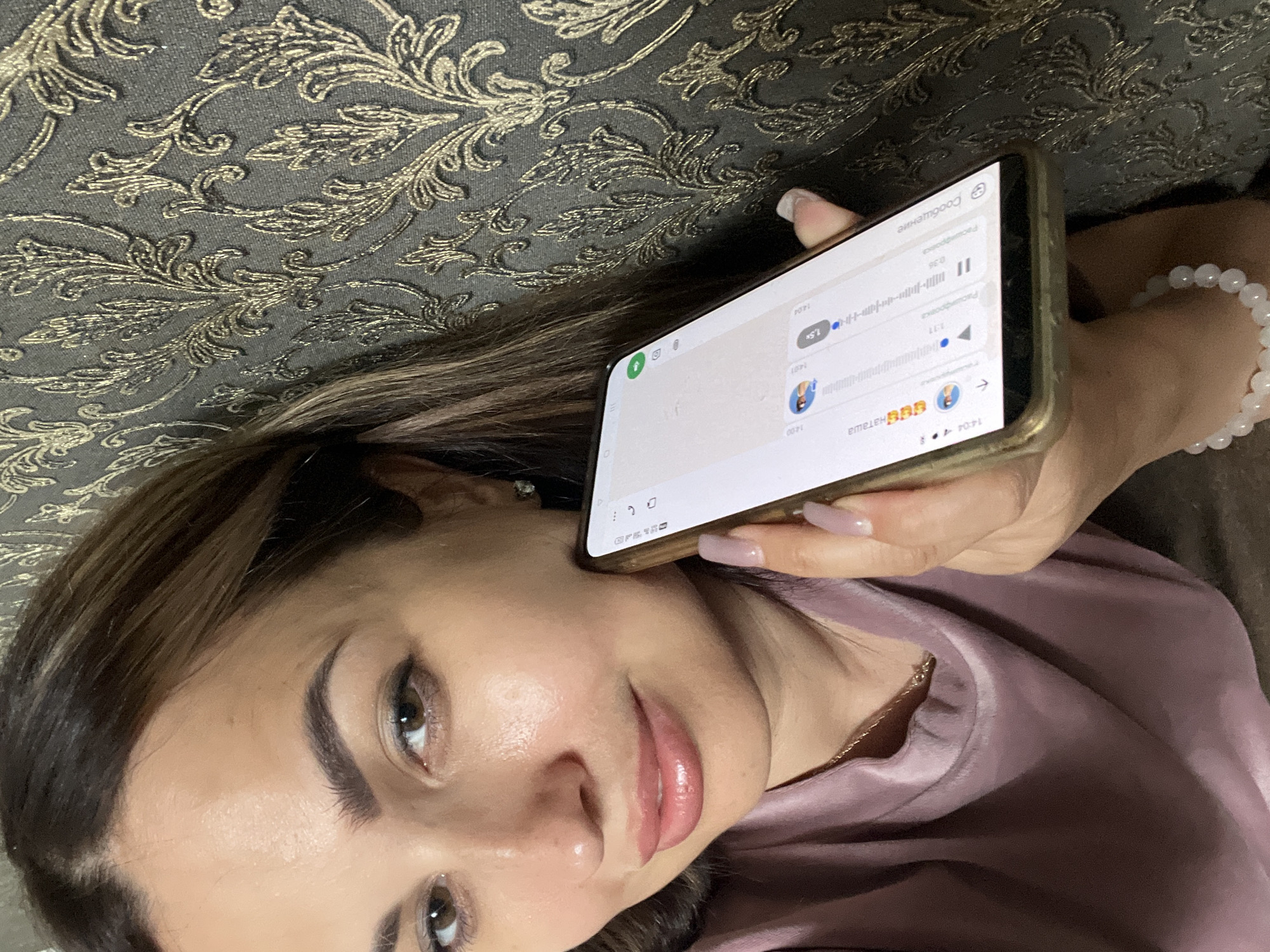 A woman with brown hair and makeup is holding a smartphone to her ear. The phone screen displays a messaging application with chat bubbles and a waveform, indicating an audio message. The woman is wearing a pinkish-purple shirt and a necklace. The background is a textured wall with a gold floral pattern. The lighting is soft, and the overall scene appears to be indoors.