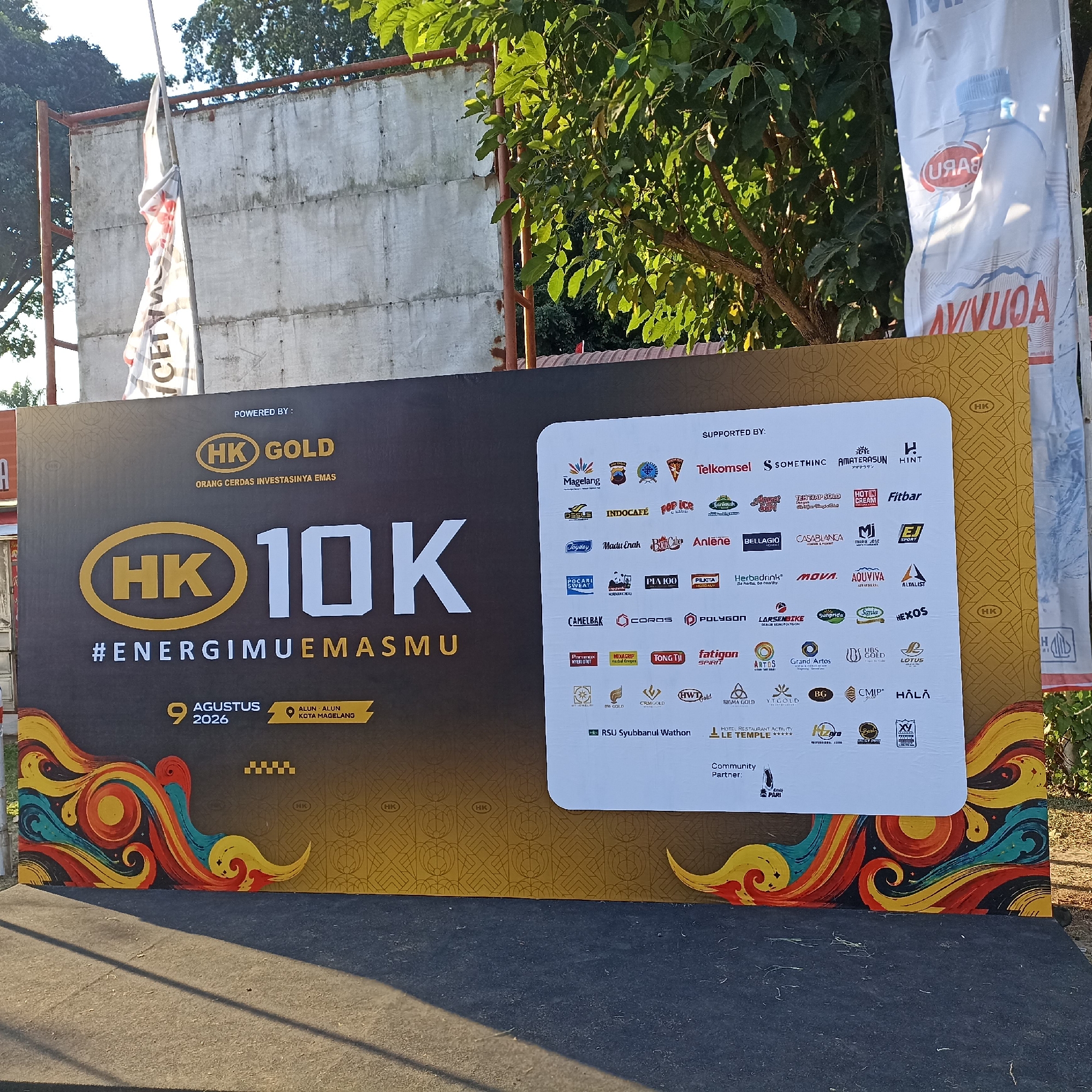 The image shows a large banner for an event called "HK GOLD 10K #ENERGIEMUEMASMU". The banner is dark gray with gold and white lettering. The date "9 AGUSTUS 2026" is displayed, along with the location "ALUN-ALUN KOTA MAGELANG". The banner also features colorful, abstract swirling designs on the left side. The right side of the banner lists "SUPPORTED BY:" and displays numerous logos of various companies and organizations, including Telkomsel, Indocafe, Pocari Sweat, and many others. Below the list of sponsors, the text "Community Partner:" is visible with a few more logos. The banner is set up outdoors, likely in a public space, as suggested by the presence of trees and a concrete wall in the background, and a black carpet laid out in front of it, possibly for attendees to walk on. The lighting suggests it is daytime, with sunlight casting shadows.