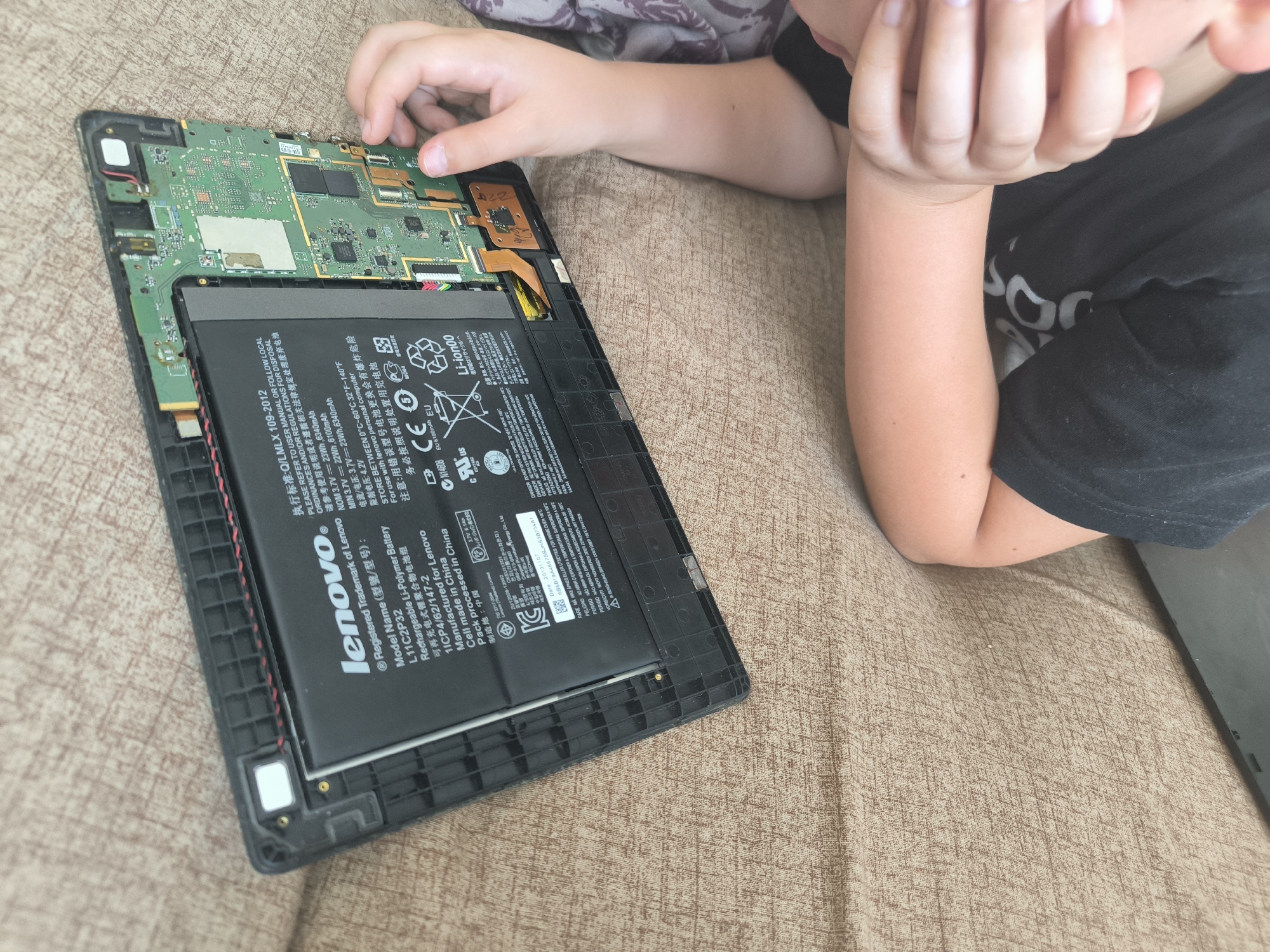 The image shows a disassembled Lenovo tablet with its internal components exposed. A person's hands and arm are visible in the upper right quadrant of the image, interacting with the tablet's circuit board. The tablet is placed on a textured, brown fabric surface, possibly a couch or blanket. The scene is not indicative of any specific location in Chișinău, Moldova, as there are no identifiable landmarks or contextual clues. The focus is on the technological components and the interaction with them.
