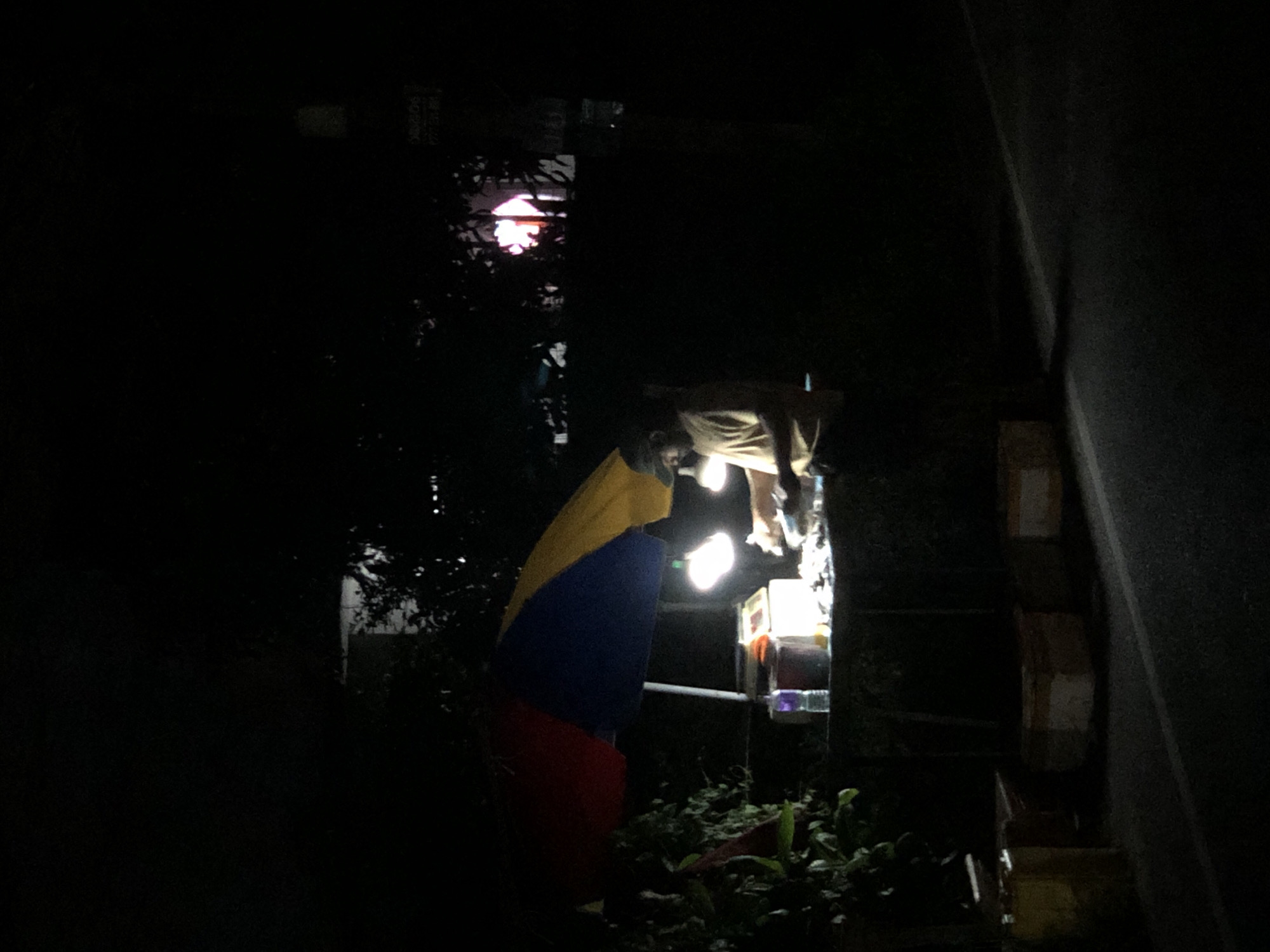 An outdoor scene at night features a man preparing food at a roadside stall. The stall is covered by a colorful umbrella, red, blue, and yellow, and illuminated by two overhead lights. The man, wearing a light-colored t-shirt and a cap, is standing behind the stall, engaged in his work. Various items, including bottles and containers, are visible on the stall. In the foreground, a paved road runs alongside the stall, with some boxes or crates placed on the side of the road. The background is dark and filled with trees, with a glimpse of a building and lights in the distance. The overall ambiance is that of a typical street food vendor operating after dark.