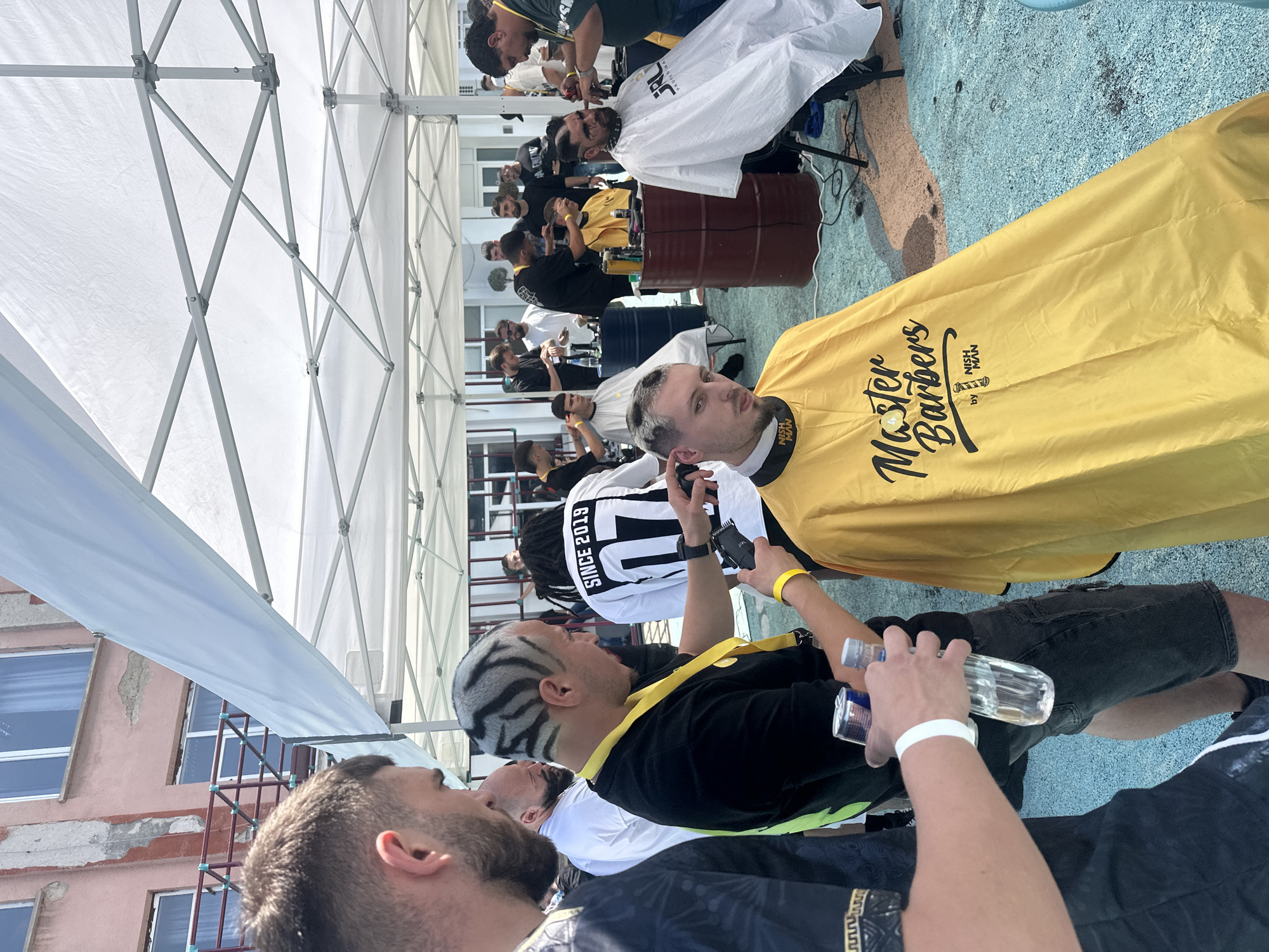 The image captures an outdoor barber event or competition taking place under several white pop-up canopy tents in Sofia, Bulgaria. Approximately 15-20 adult males are present, functioning as barbers, clients, and observers.
In the foreground, a client with a partially buzzed haircut, mustache, and soul patch is seated, wearing a yellow barber's cape that reads "Master Barbers by NISH MAN." To his left, a barber with a distinct black and white zebra-striped pattern shaved into his hair, wearing a black t-shirt and a yellow lanyard, is actively using a hair clipper on the client's head. Another man, partially visible on the far left with a beard and dark patterned shirt, holds a Red Bull can and a plastic water bottle, sporting a white wristband.
Further to the right, another client with a beard and dark facial markings is seated, draped in a white cape displaying "SRL PROFESSIONAL." A barber in a dark green shirt and yellow lanyard attends to his hair.
Throughout the scene, multiple barber stations are established, featuring client chairs and barbers working on various clients. Dark blue and brown industrial barrels are repurposed as stands for barber tools. One individual in the background wears a white t-shirt with "SINCE 2019" and a large "7" on the back.
The ground surface is a patchy, textured mix of light blue and reddish-brown, indicating a worn outdoor pavement or court. In the background, beyond the tents, a multi-story building with a weathered pink/peach facade and white-framed windows is visible, along with another lighter-colored building. The event occurs under bright daylight conditions.