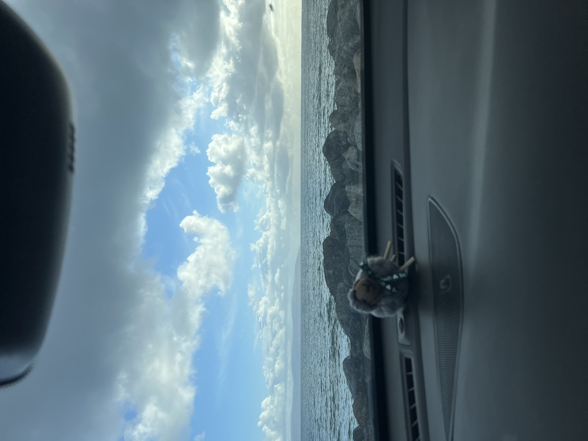 The image depicts a coastal view from inside a vehicle, likely a car. The landscape consists of a body of water with a rocky shoreline visible on the right side of the frame. The sky overhead is filled with a mix of cumulus clouds, varying in shades from bright white to dark grey. Part of the car's interior, including the dashboard and a rearview mirror, are visible, and there's a small stuffed animal toy attached to the dashboard.
Due to the lack of distinguishable landmarks, the exact location within Galway, Ireland, cannot be precisely determined, but the image suggests a coastal area.