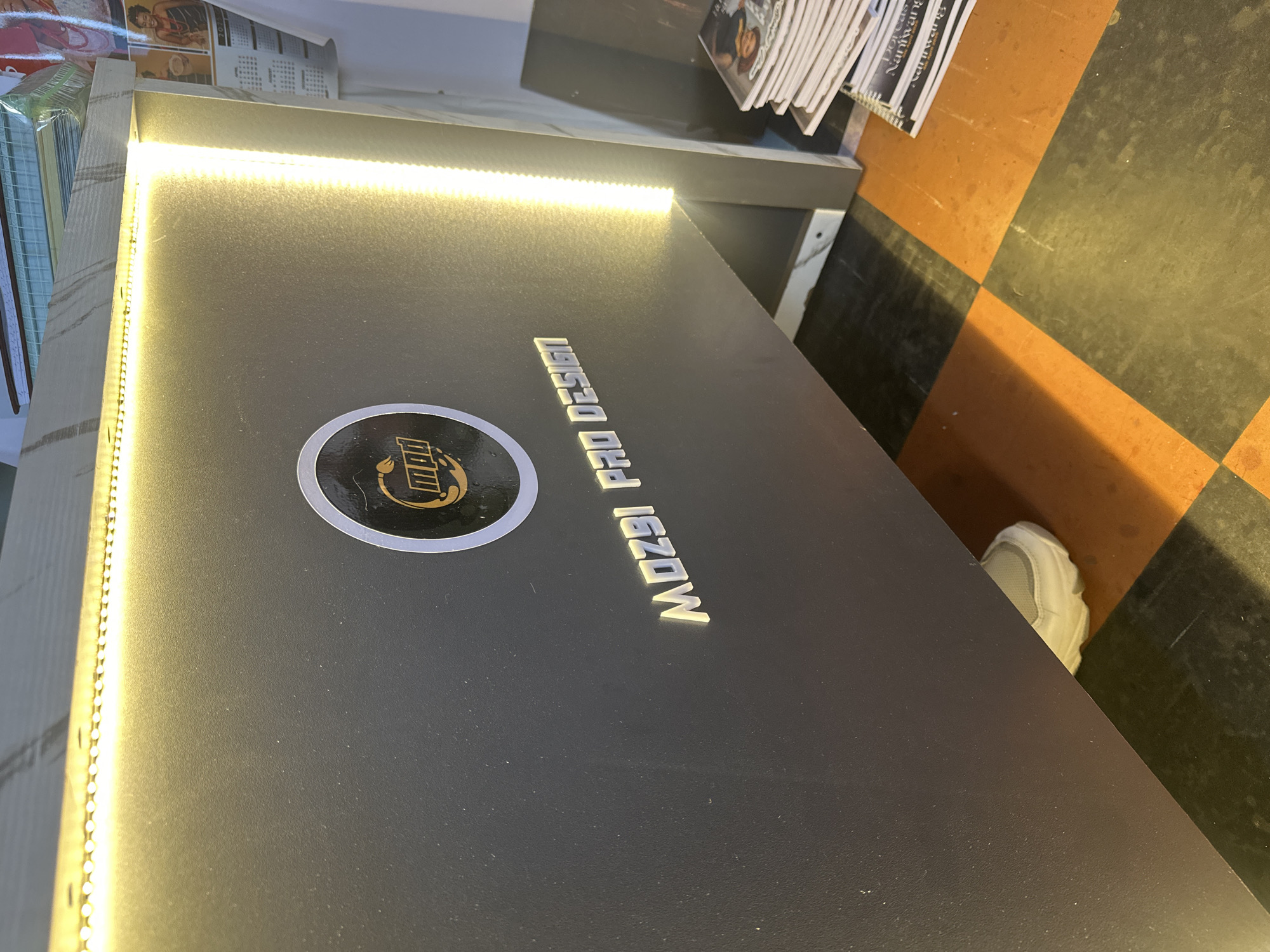 The image depicts a close-up view of a dark gray surface with a luminous LED strip along its edge. A circular logo with the word "MP" and an icon is visible on the surface, along with the text "MOZ9I PRO DESIGN". In the background, there are stacks of books or magazines. A white shoe is visible near a floor with alternating dark and light squares in the background. The location is unknown, as Gura Top is in Nigeria, and there is no indication of this in the image.