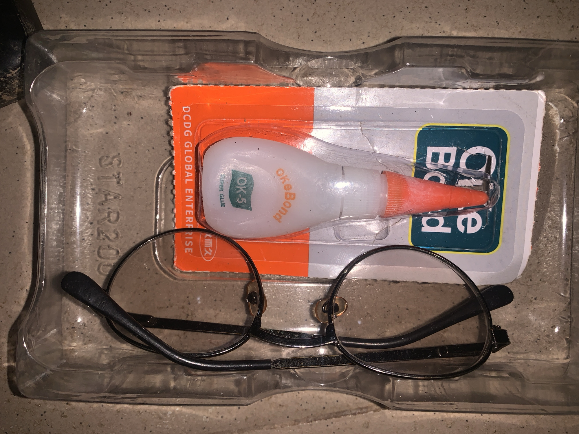 The image shows a pair of black-rimmed glasses placed inside a clear plastic container with a card featuring a tube of "OK Bond" super glue. The glasses are lying on their side, with the lenses facing upwards. The glue tube is white with an orange tip and has the brand name "OK Bond" and "OK-5 SUPER GLUE" printed on it. The container and the glue packaging are sitting on a rough, textured surface that appears to be concrete or stone. The background is dark and out of focus, suggesting the image was taken in low light conditions. There are no people visible in the image. The location is unknown.
