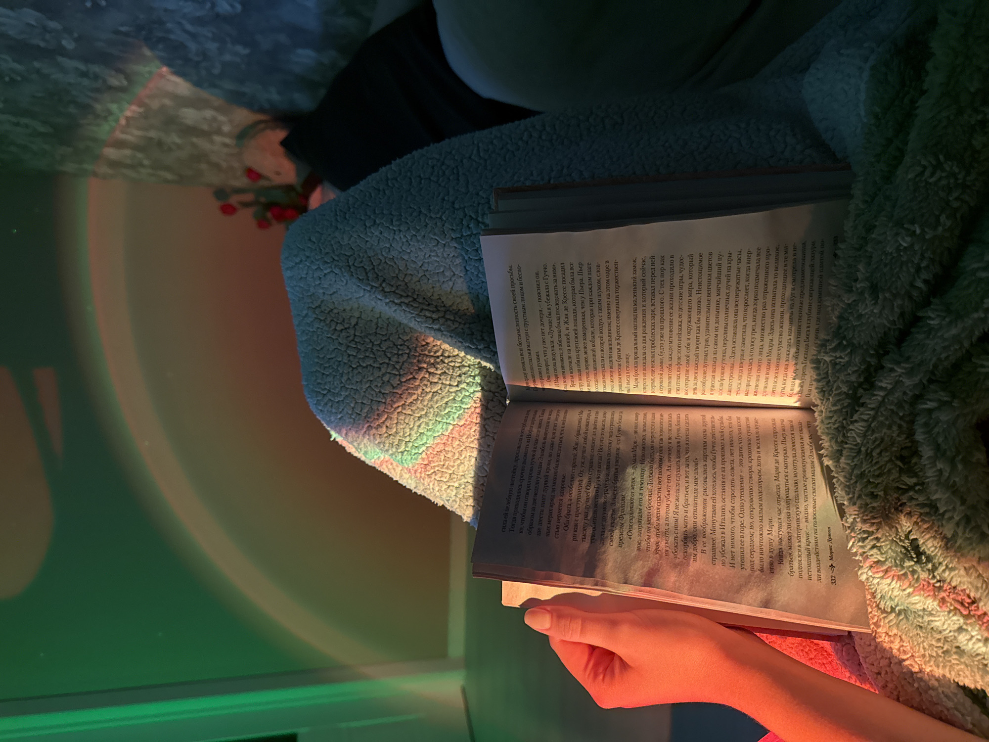 Here is a description of the image:
**Subjects**: The image features a person holding an open book and is wrapped in a textured blanket or throw.
**Objects**: The primary objects are a book with visible text and the blanket. There is also a glimpse of a small red object (possibly berries or small flowers) and an undefined dark object partially visible in the top left of the frame. Additionally, there's a subtle green and orange light cast on the scene.
**Environment**: The environment appears to be indoors. The lighting is soft and artificial, with green and orange light creating a warm, cozy ambiance.
**Details**: The book appears to be open to a page with text in a language other than English (possibly Russian), suggesting it may be a novel or a work of fiction. The blanket looks soft and warm, adding to the feeling of comfort. The person's hand and arm are subtly illuminated by the artificial lights. The image is vertically oriented.