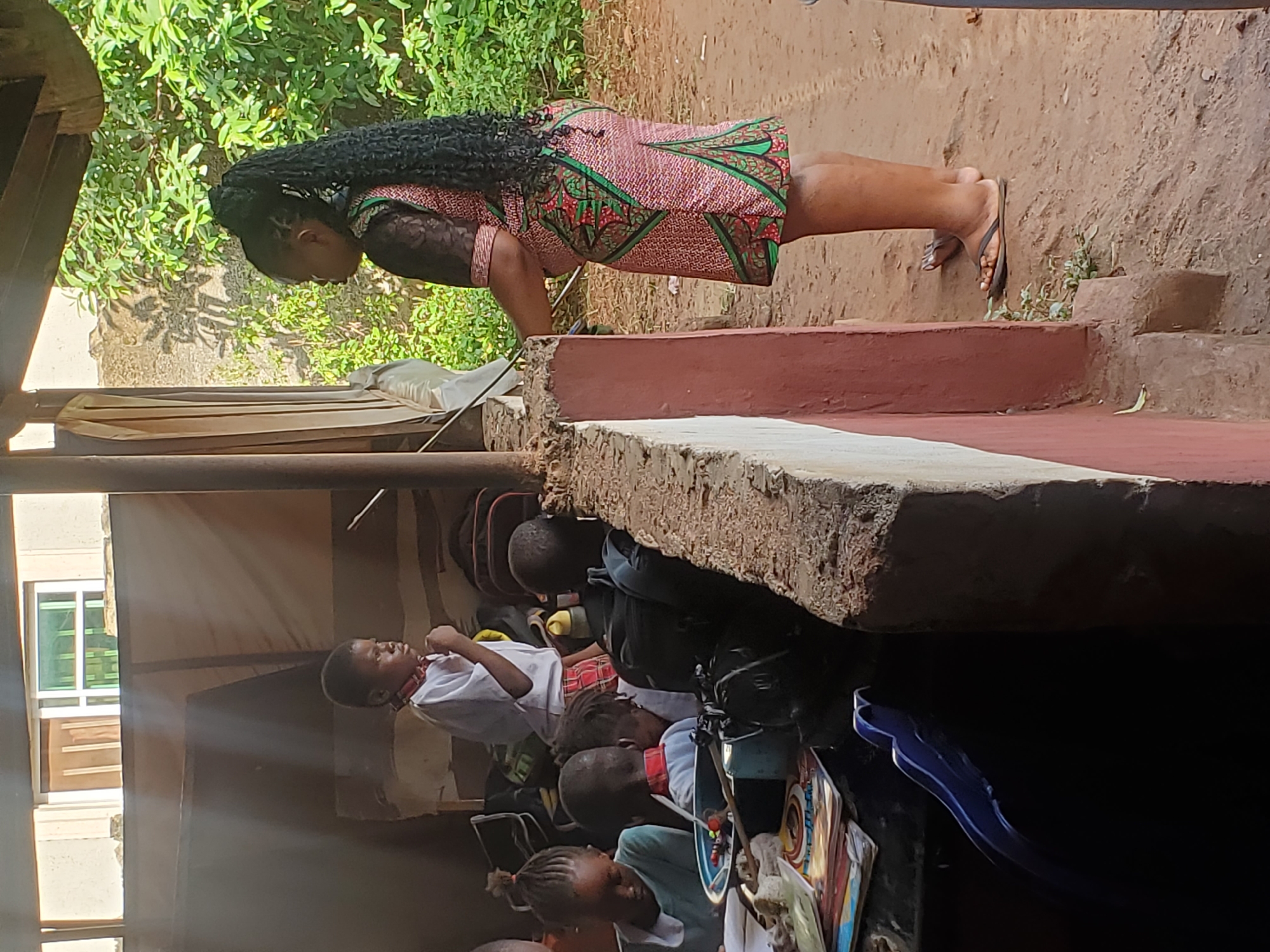 The image captures a scene both indoors and outdoors in what appears to be a less developed urban or semi-rural area. Outside, a woman with long, braided hair is standing on a raised surface, likely a step or low wall, wearing a colorful patterned dress and sandals. She is positioned next to a grassy area or vegetation.
Below her, the scene transitions to an indoor space. Several children, some wearing what seems to be school uniforms, are visible. There are bags, possibly school bags, and other objects like books and a blue utensil in the indoor space. Sunlight filters in through a window in the background. It seems the children are in some kind of classroom or learning environment. The exact city and country location are not discernible from the image.
