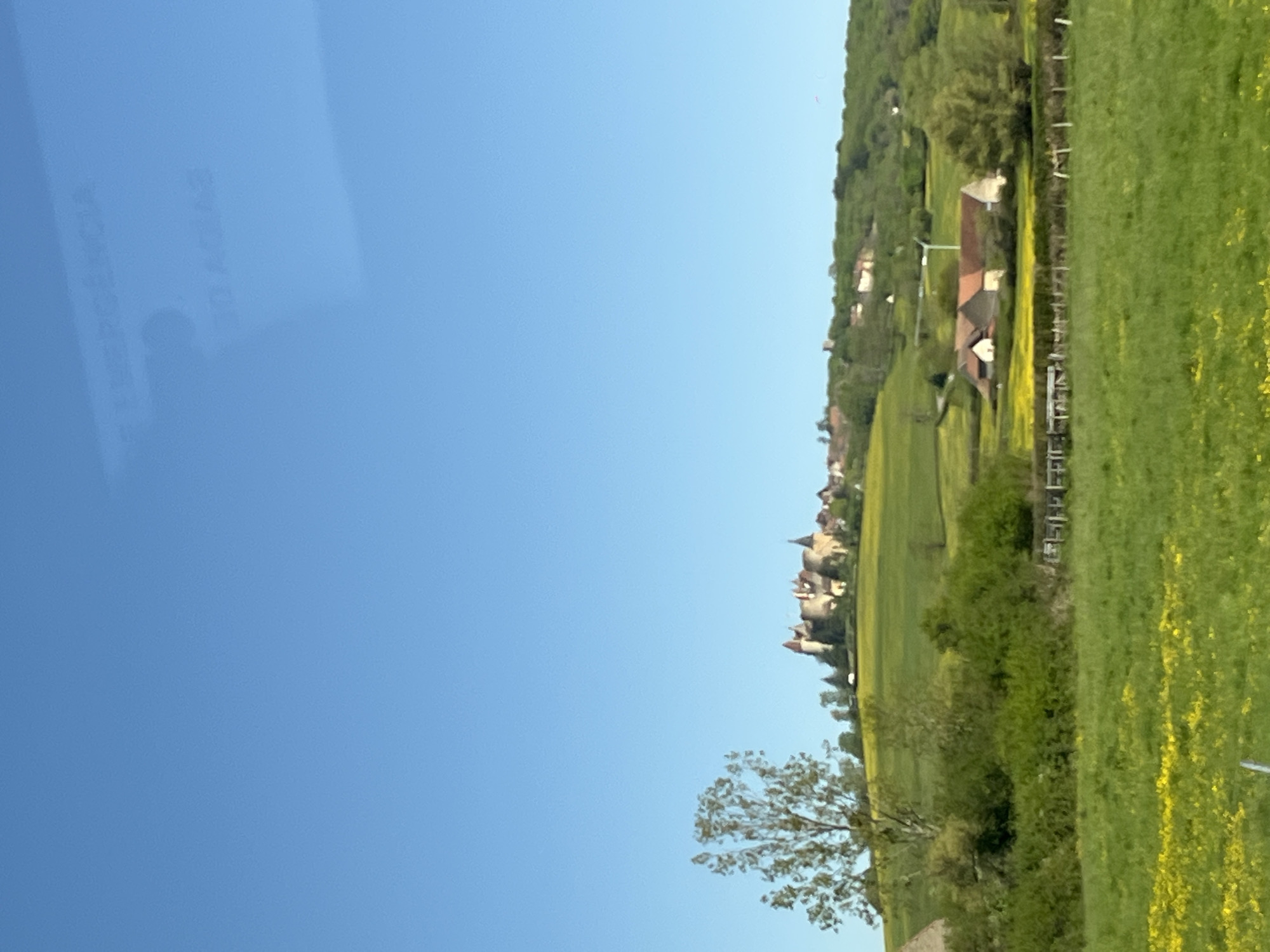 The image shows a landscape in Châteauneuf, France, featuring a clear blue sky occupying the left side of the frame. The right side presents a countryside vista, with green meadows, trees, and scattered houses dotting the terrain. In the mid-ground, a cluster of buildings resembling a medieval village or castle is visible, presumably part of Châteauneuf's historic architecture. A wooden fence runs along a field. The foreground includes some foliage and a bare tree, likely adding depth to the landscape view. A reflection, blurred, in the window indicates some words. The overall impression is a sunny day in a rural French setting.