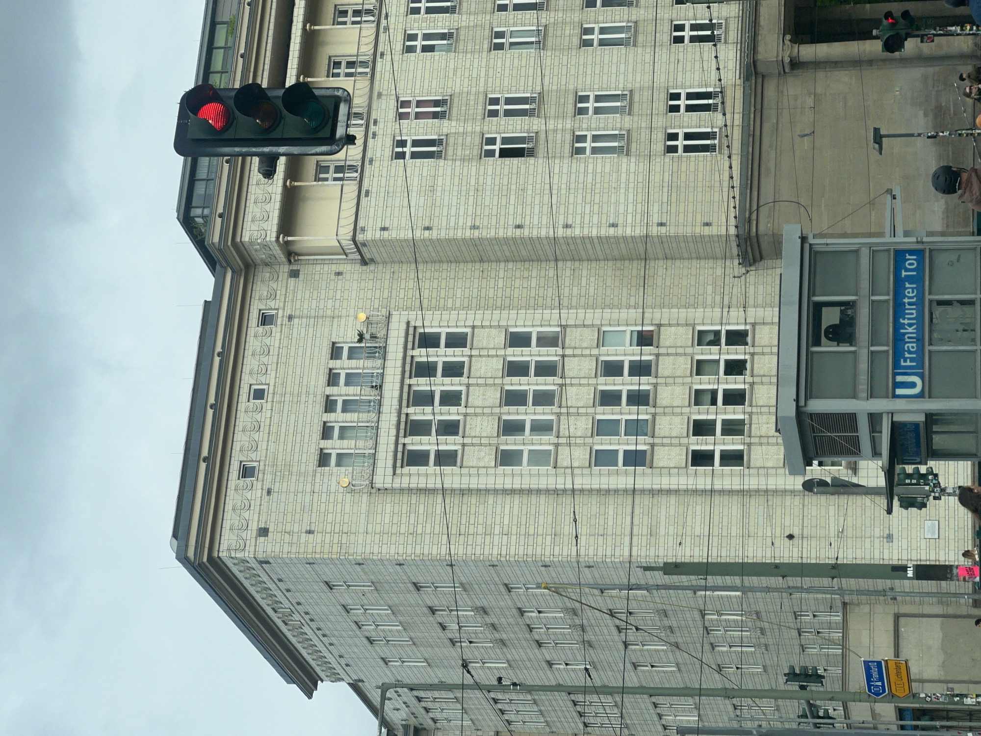 The image shows a view of Frankfurter Tor in Berlin, Germany. The architectural style of the building is prominent, featuring a light-colored facade with rows of windows. A traffic light displays a red signal. A sign for the U-Bahn station "Frankfurter Tor" is visible. The sky is overcast, suggesting a cloudy day. There are visible power lines in the foreground. A street sign indicates a route to Frankfurt/O. People are seen in the background near the station.