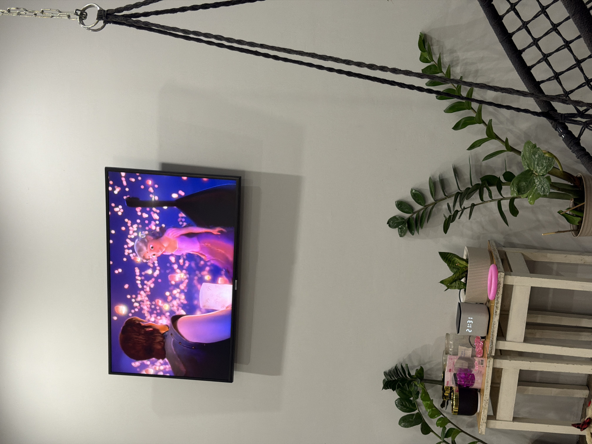 The scene presents an interior wall featuring a television playing the movie "Tangled." The foreground includes a hanging swing or hammock. Adjacent to the television is a multi-tiered shelf unit, populated with plants, a digital clock displaying "21:31," and various decorative items. The wall color is a neutral gray. The location is unknown, despite the requested inclusion of information about Chaikovsky, Russia.