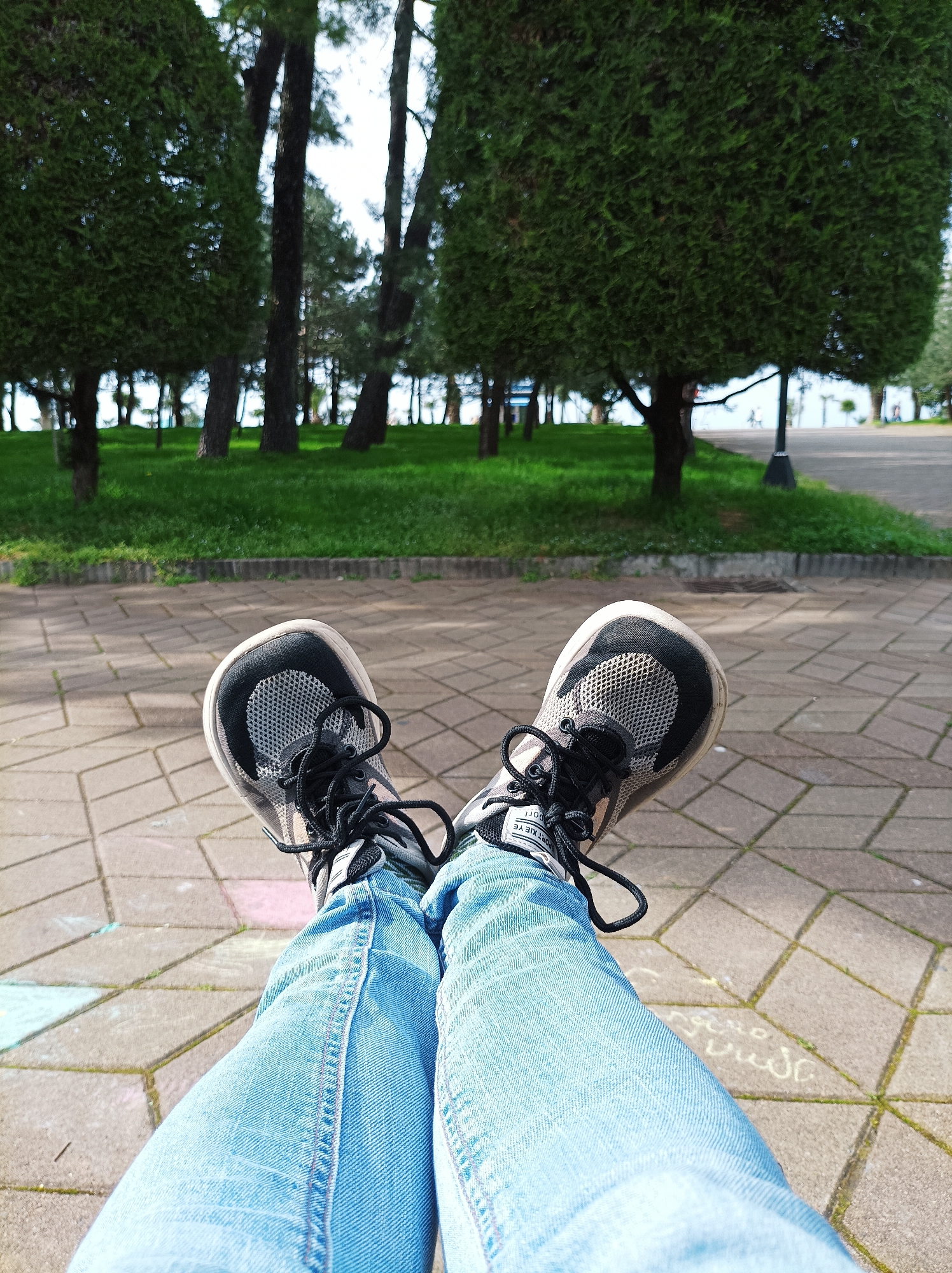 A point-of-view shot shows a person sitting with legs outstretched on a patterned concrete surface in Batumi, Georgia, likely in a park setting. The person is wearing light blue denim jeans and a pair of black and grey running shoes with black laces. The background features a grassy area with trees and a paved walkway. Several neatly trimmed, square-shaped trees are visible, and other, taller trees are in the background. In the lower right of the frame, there is white writing in cursive script on the pavement next to the person's leg, suggesting the area may be a space for recreational activities like drawing with chalk. A lamppost and glimpses of a walkway with people in the distance are seen behind the grass and trees.