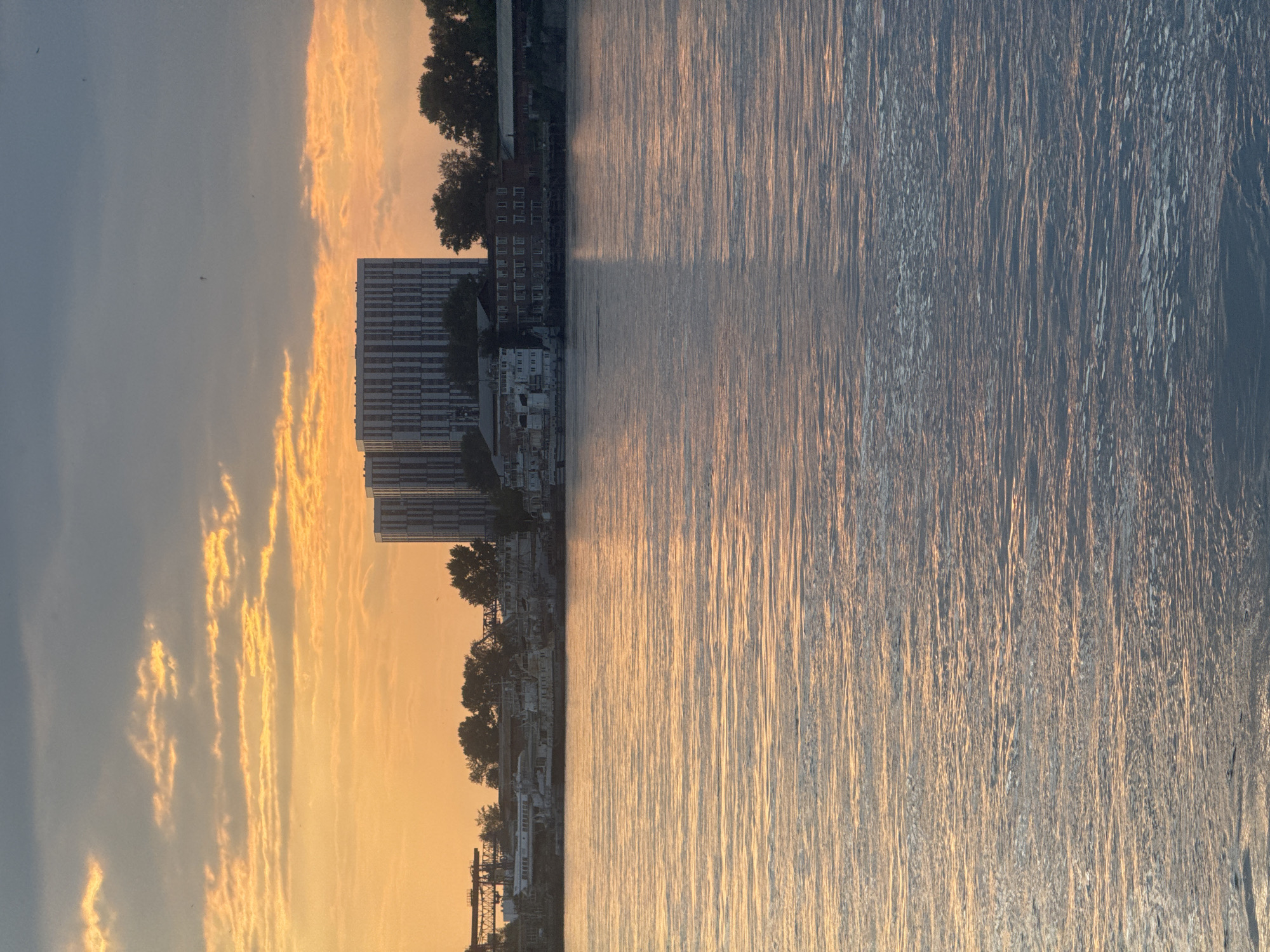 The image captures a serene sunset over the Angara River in Irkutsk, Russia. The sky is a canvas of warm oranges and yellows, painted with streaks of clouds against a darkening blue backdrop. Two imposing, modern high-rise buildings dominate the middle ground, their vertical lines contrasting with the horizontal expanse of the river. Along the riverbank, a collection of older buildings and industrial structures, including several boats docked along the shore, are visible. The water itself reflects the colors of the sunset, creating a shimmering, undulating surface that fills the foreground and mid-ground of the image. The overall atmosphere is tranquil and reflective, showcasing the confluence of urban development and natural beauty at dusk.
