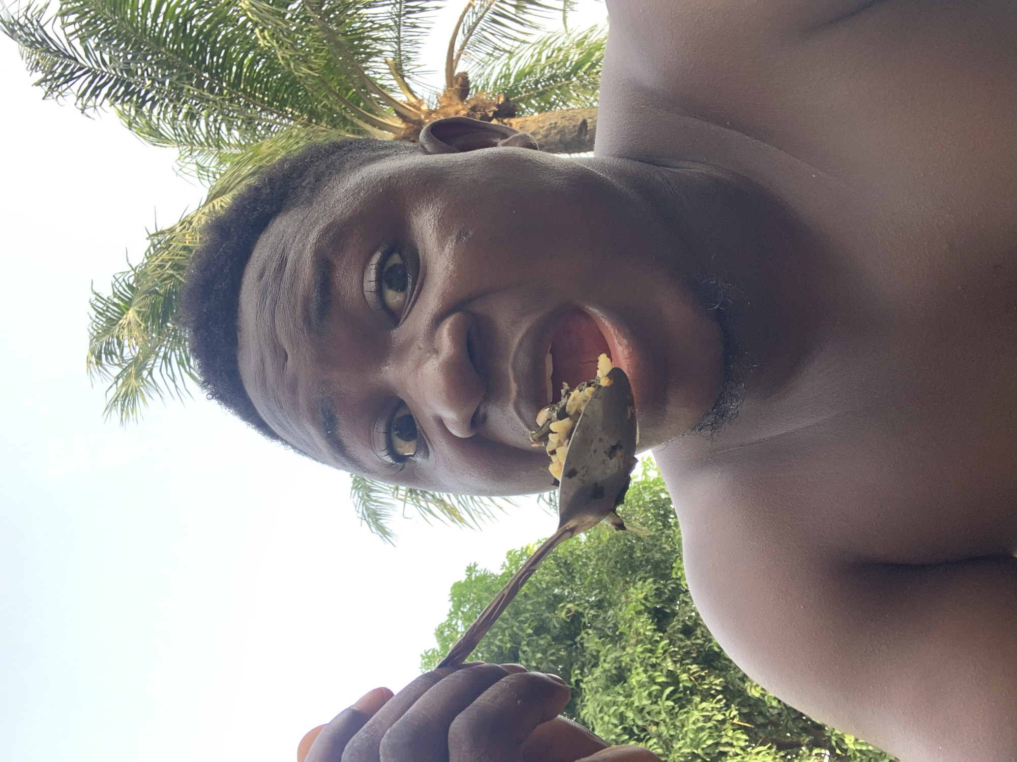 A close-up, low-angle portrait captures a young person with dark skin and short, dark hair, seemingly taken from below, looking up. The individual, with a bare upper body, holds a spoon laden with food, appearing to be rice mixed with green vegetables, towards their open mouth. Their left eye (viewer's right) is wide open and looking directly at the camera, while their right eye (viewer's left) is squinted, conveying a playful or cheeky expression.
The background features a bright, clear sky dominating the upper portion of the frame. Lush green foliage, including the fronds of a palm tree, is visible behind and above the person's head, suggesting an outdoor setting during the daytime. The lighting is bright and natural. The image focuses intently on the person's face, mouth, and the food on the spoon, with details of their skin texture clearly visible. No visible text is present in the image. The scene suggests a casual moment of eating outdoors, possibly in a warm, tropical climate like Akwanga, Nigeria.