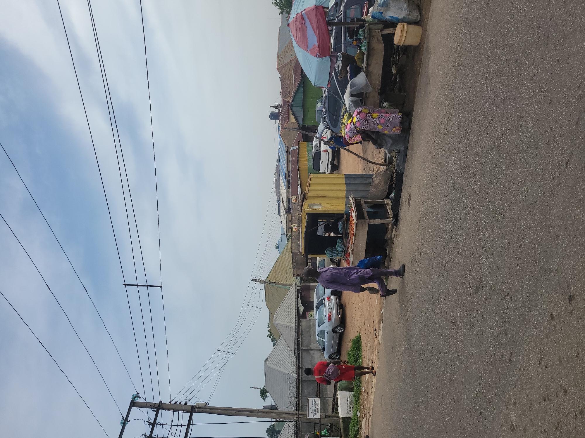 The image depicts a street scene in Jos, Nigeria, under an overcast sky. In the foreground, a paved road stretches across the frame. To the left, a person in a red shirt and shorts walks away from the camera, carrying a backpack. Further back, a man in a purple traditional outfit walks towards the right, holding a dark bag.
On the right side of the road, a makeshift market stall is set up. Several people are attending to goods displayed on tables. Two women are visible, one in a colorful patterned dress, interacting with produce. A partial view of a market umbrella, with red and blue sections, is present. Various vehicles, including silver sedans and a white pickup truck, are parked in the background, along with several market stalls and residential buildings with corrugated metal roofs. Electrical poles and overhead wires are prominent throughout the scene, indicating urban infrastructure. The ground around the market stall is dirt and debris.