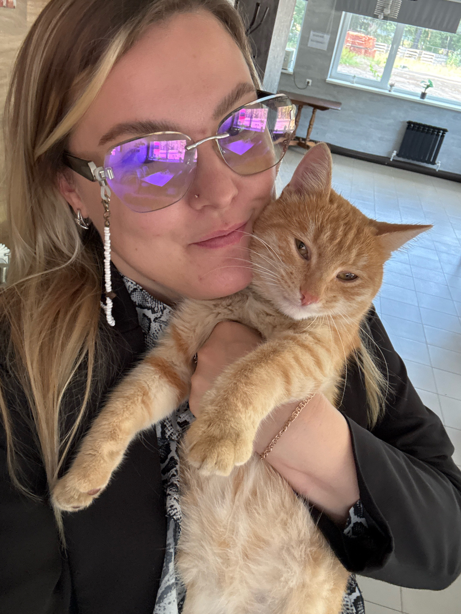 An individual, identified as a Caucasian female with blonde hair, is holding an orange tabby cat. She is wearing glasses with purple lenses, a black blazer over a patterned shirt, and earrings. A beaded chain hangs from her glasses. The cat appears comfortable and is being held close to the individual's face. The background suggests an indoor location, possibly a public building or waiting area, with a window showing an outdoor scene with greenery and structures. There are radiators and furniture visible. The floor is tiled. The image was taken in Noviy, Russia.
