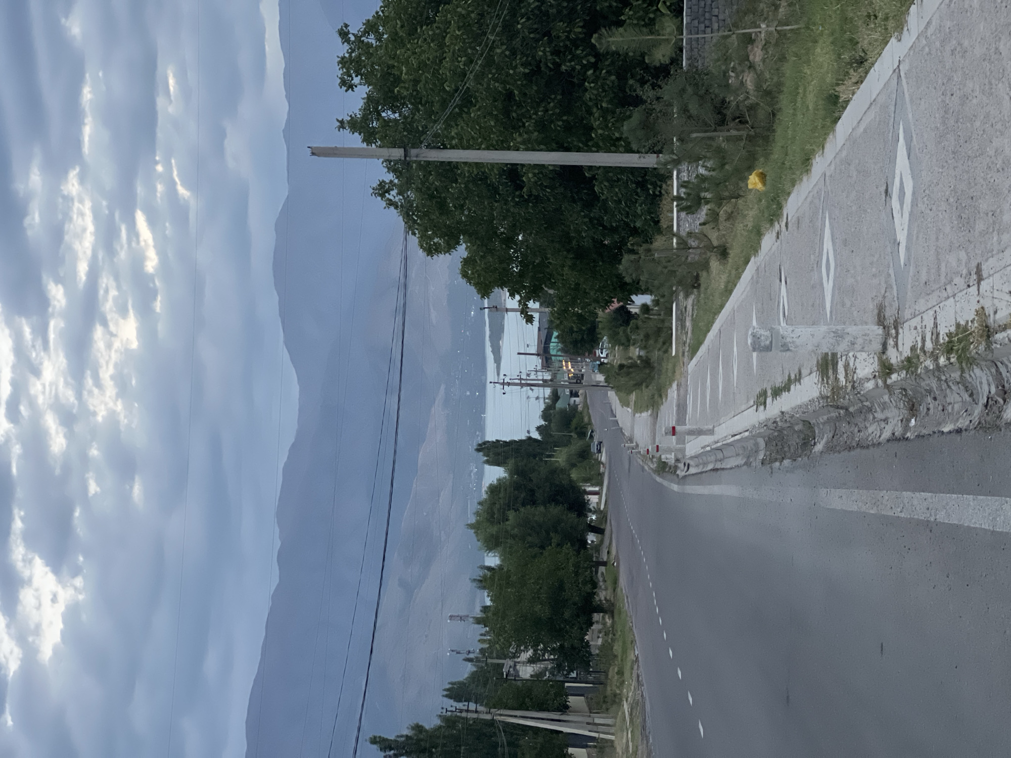 This is a vertical, eye-level, outdoor shot of a road and sidewalk in Yusuphana, Uzbekistan. The sky is overcast with muted gray clouds. In the foreground, a paved road curves to the left, marked with white dashed lines and a solid white line. To the right of the road is a concrete sidewalk with square decorative patterns. There is a drainage ditch along the edge of the road. A few small trees and bushes line the sidewalk. In the midground, there are more trees, some taller, and several utility poles with wires. A small dark-colored car is visible on the road. In the background, a body of water, likely a lake or reservoir, is visible, bordered by distant, hazy mountains. The overall scene depicts a somewhat subdued, possibly late afternoon or early morning atmosphere.