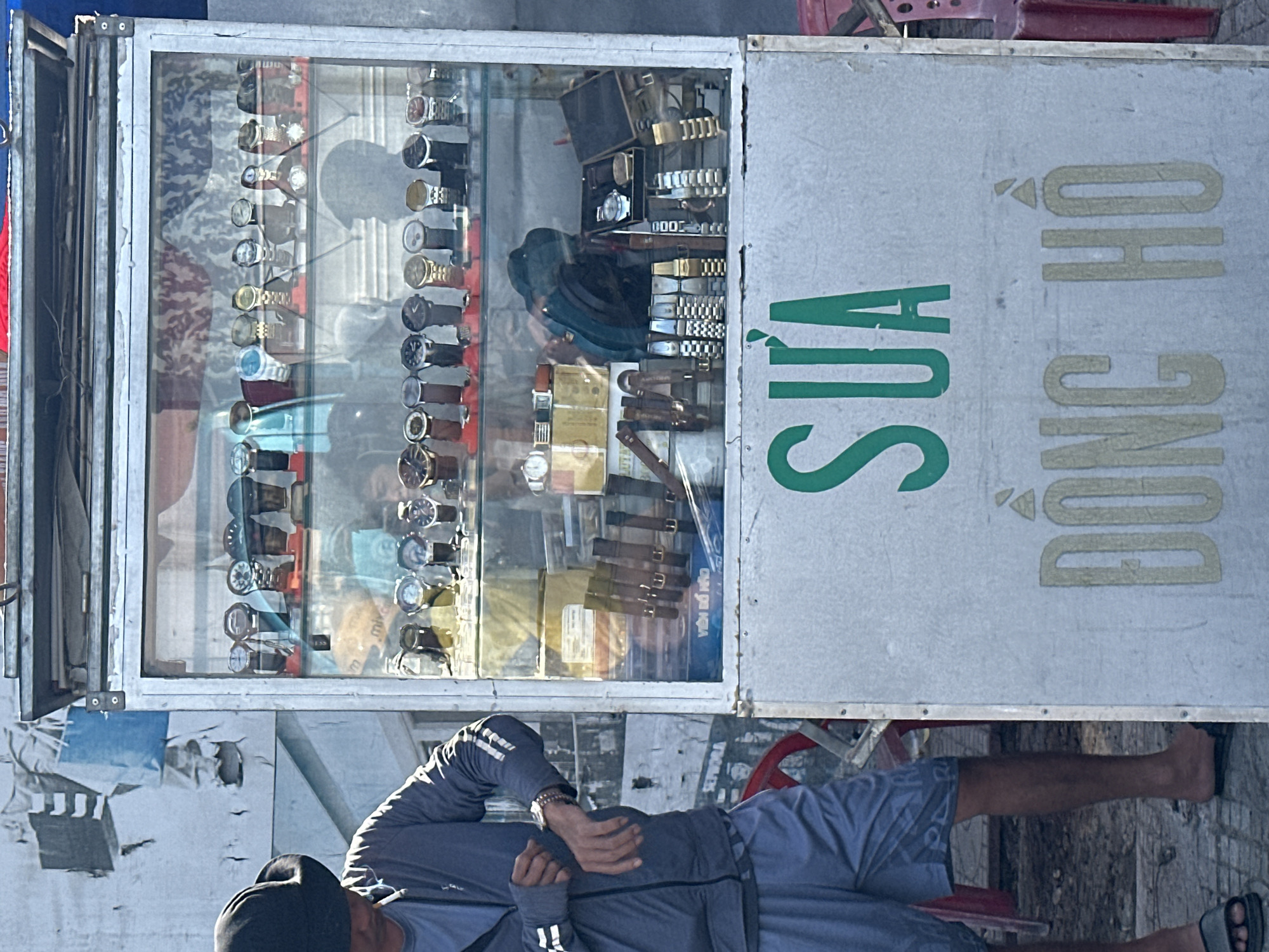 A man wearing a black hat, gray zip-up jacket, and gray shorts stands to the left of a glass display case filled with watches. He is adjusting his left sleeve, revealing a watch on his left wrist. The display case is positioned on a stand with a sign that reads "SUA" in green lettering at the top and "DONG HO" in beige lettering below. The background includes a white wall with peeling posters and a red chair. The scene appears to be outdoors, likely on a street in Bac Nha Trang Ward, Vietnam, given the signage.