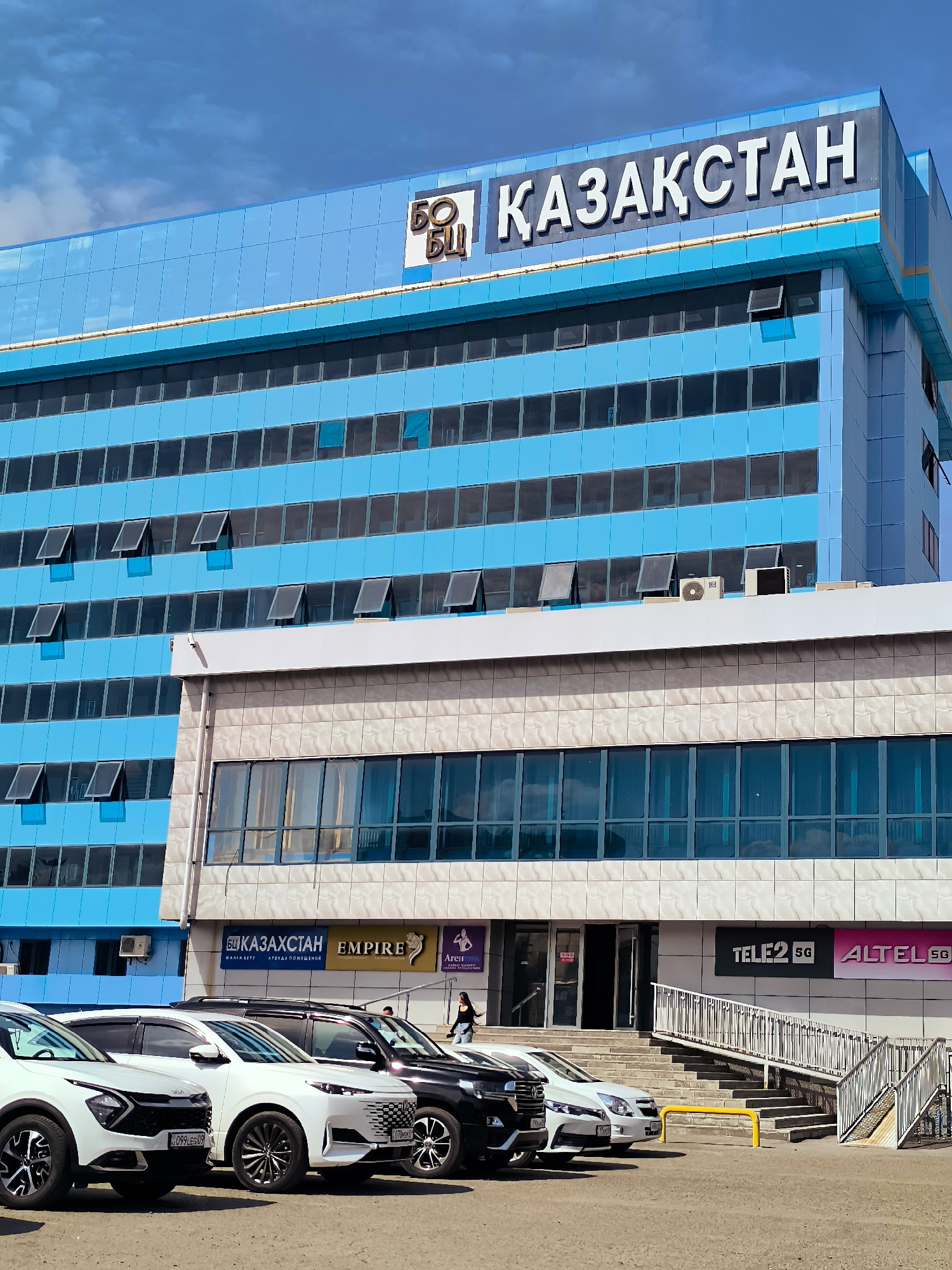 The image shows a modern, blue multi-story building with the text "ҚАЗАҚСТАН" (Kazakhstan) on top. The building features numerous windows, many of which are partially open. Below the blue section, there is a section with beige tiled walls and darker windows. Several cars are parked in front of the building, including white SUVs and a black SUV. Two women are visible near the entrance of the building, one walking away from the camera and the other walking towards the building. Signage on the lower part of the building includes "БО БИ", "ҚАЗАҚСТАН", "EMPIRE", "Агент", "TELE2 5G", and "ALTEL 5G". The scene is outdoors on a sunny day with a cloudy sky.