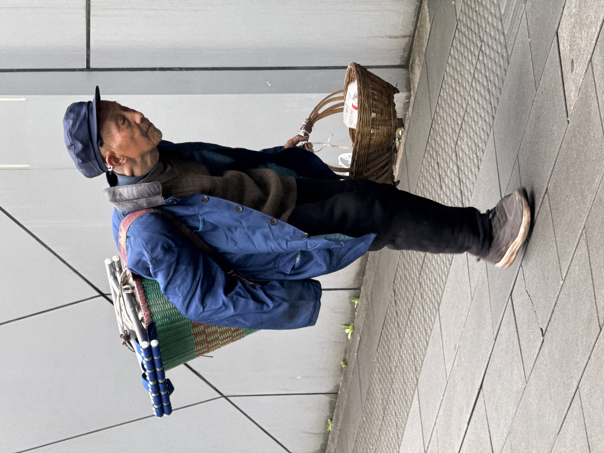 A man in a blue cap, brown sweater, blue jacket, and black pants stands on a gray tiled sidewalk next to a gray wall. He has a basket on his back and another wicker basket he is holding. The location is Wulong District, China.