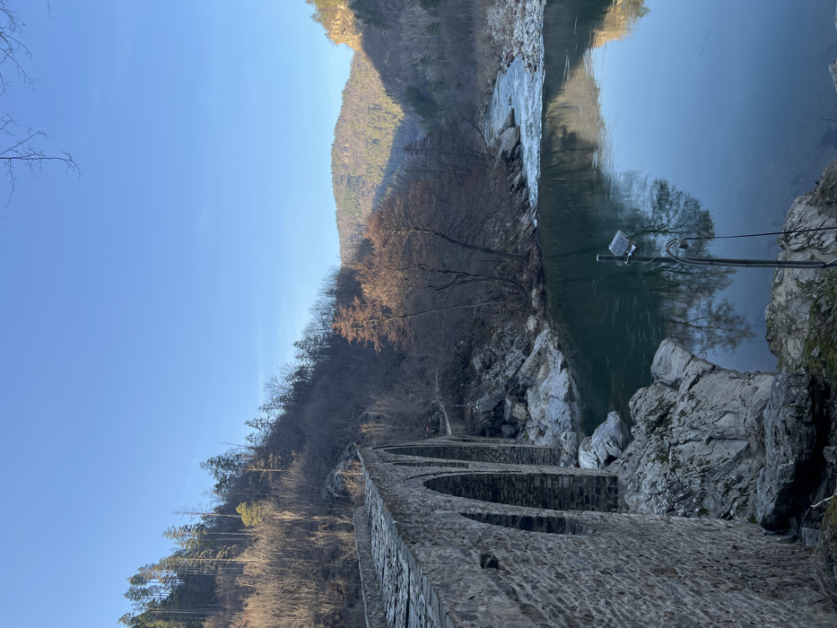 The image displays the Devil's Bridge (Dyavolski most) in Diadovtsi, Bulgaria, crossing a river within a steep, forested valley. The multi-arched stone bridge, constructed from rough-hewn blocks with a cobblestone surface, extends from the lower left of the frame towards the center. The river flows beneath the bridge from the background, presenting dark green-blue water that reflects the clear blue sky and surrounding vegetation. Visible upstream of the bridge are areas of white rapids.
The riverbanks are characterized by numerous large rocks and boulders. Steep hillsides rise on both sides of the river, densely covered with a mix of deciduous trees, many bare, and evergreen coniferous trees, indicating a season outside of full summer foliage. Distant mountains, also tree-covered, are visible in the background under a clear, bright blue sky. Sunlight illuminates the scene from the right, highlighting the river's surface, parts of the bridge, and portions of the distant hills, while the left hillside remains partially shadowed. In the lower right foreground, a metal pole stands on the riverbank, supporting a small solar panel and an attached camera or sensor device. No individuals are present in the scene.