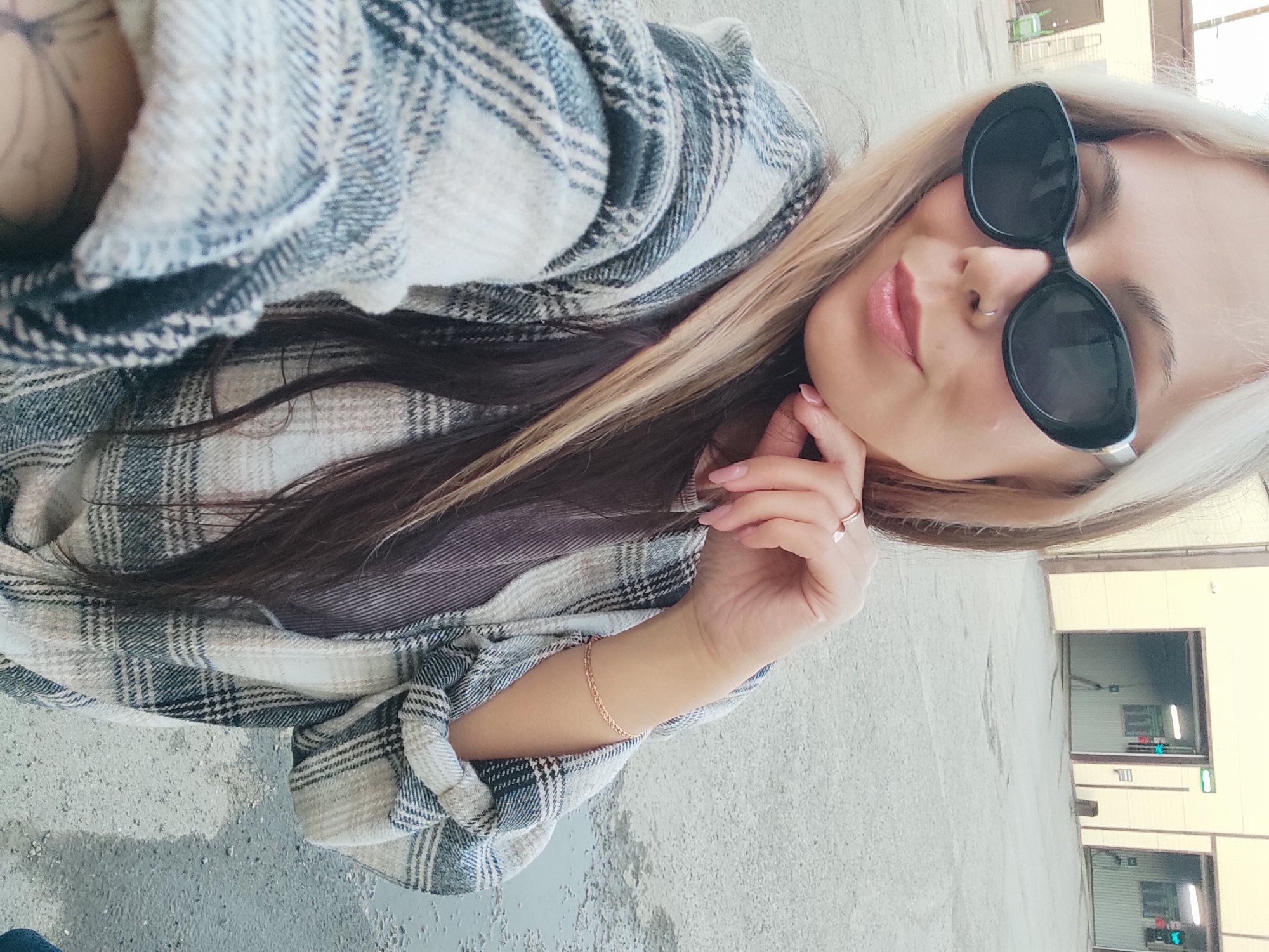 A young woman is captured in a selfie-style portrait, with her head tilted, giving the impression of a horizontal pose. She is situated outdoors in what appears to be Chaikovsky, Russia. The woman has long, flowing hair, styled with blonde highlights or a two-toned effect, contrasting with darker strands underneath. Her eyes are obscured by large, black oval-shaped sunglasses. A small, silver hoop nose piercing is visible on her nostril. Her lips are adorned with soft pink lipstick, and her fingernails are painted a light, neutral pink.
She is wearing a plaid shirt in shades of white, grey, and dark blue or black, with the sleeves rolled up. A thin, gold-toned chain bracelet is visible on her right wrist, and a delicate ring is worn on one of her fingers. Her right hand is gently positioned near her chin, suggesting a casual pose.
The background features a light grey, possibly concrete or asphalt, paved surface, showing some wear and lighter patches. In the mid-distance, several light-colored, plain buildings with dark, rectangular openings, resembling garage doors or bay entrances, line the area. The overall lighting suggests it is daytime with clear weather conditions. The woman's partially visible expression indicates a relaxed and potentially content mood, with a slight smile evident beneath her sunglasses. No discernible text is visible in the image.