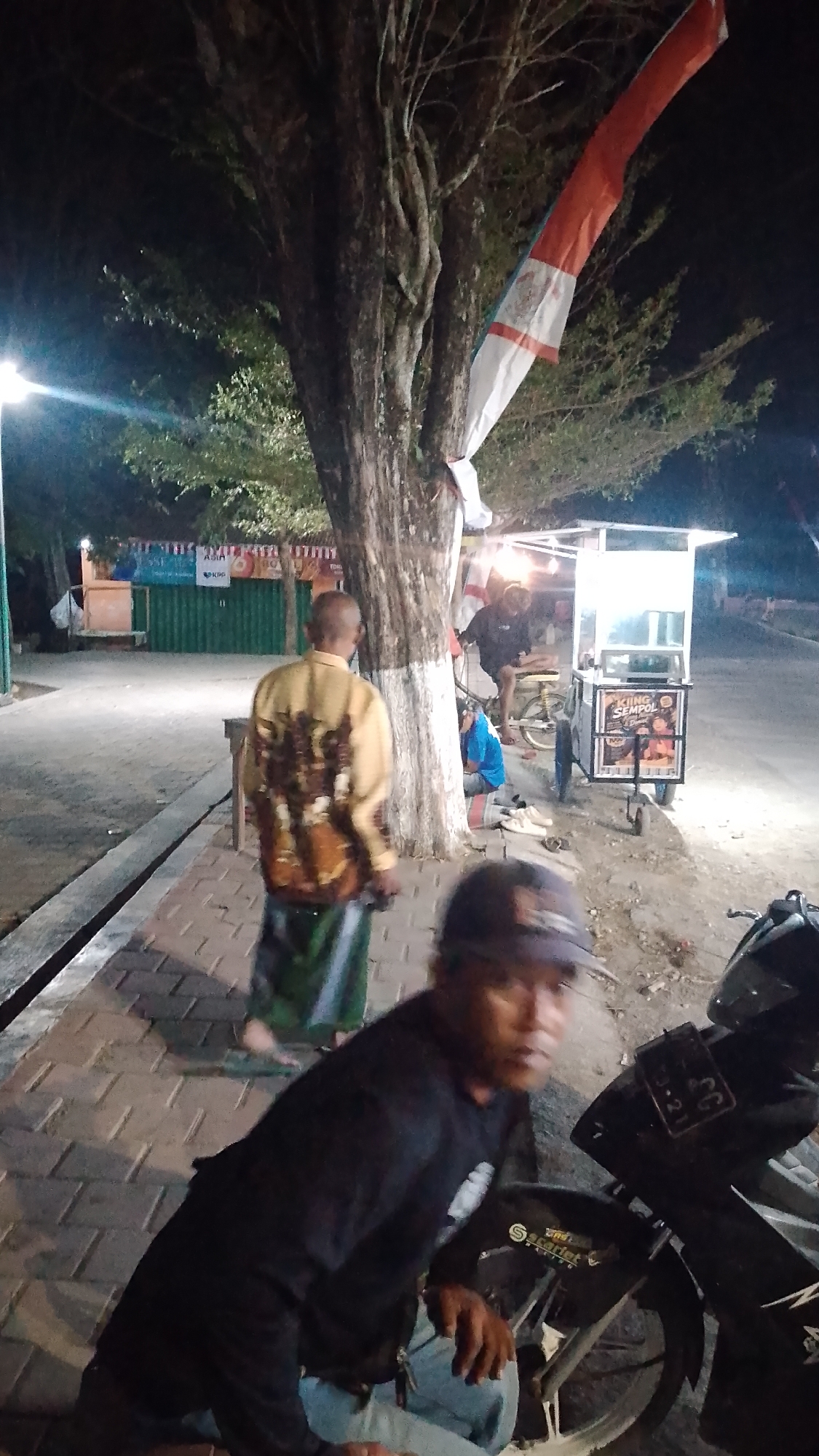 This is a nighttime photograph taken in the city of Karanglangu, Indonesia. The scene depicts an outdoor area with a large tree in the foreground, adorned with a red and white flag. A man wearing a yellow shirt with a patterned back and a green sarong is walking away from the camera, on a brick-paved walkway. Further in the background, a food cart is visible, with people around it. A motorcycle is parked on the right side of the image, with a man wearing a black shirt and a baseball cap crouching beside it, looking towards the camera. The overall lighting suggests it is after dark, with artificial lights illuminating the area.