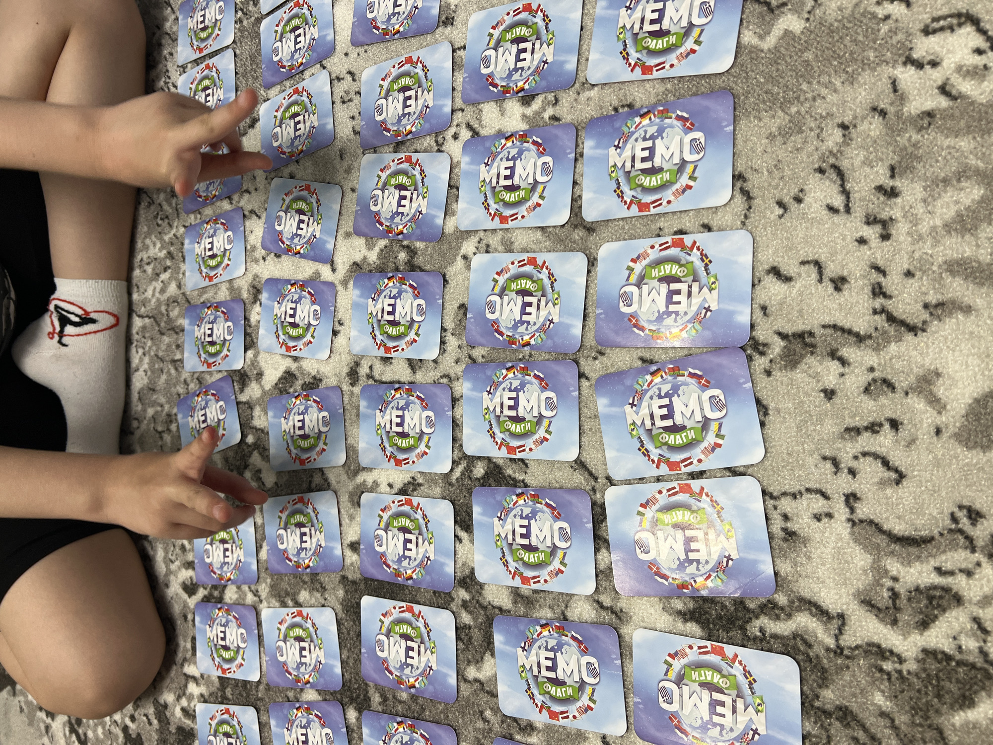 The image depicts a child playing a matching card game on a gray patterned carpet. The cards are arranged in rows and columns. Each card features the word "MEMO" and the word "FLAGS", surrounded by a circle of various national flags. The child's hands are visible, suggesting they are in the process of turning over cards. The child is wearing black shorts and white socks with a bird design. The game is laid out on a carpet inside of a home. The location of Chaikovsky, Russia, is not discernible from the image.