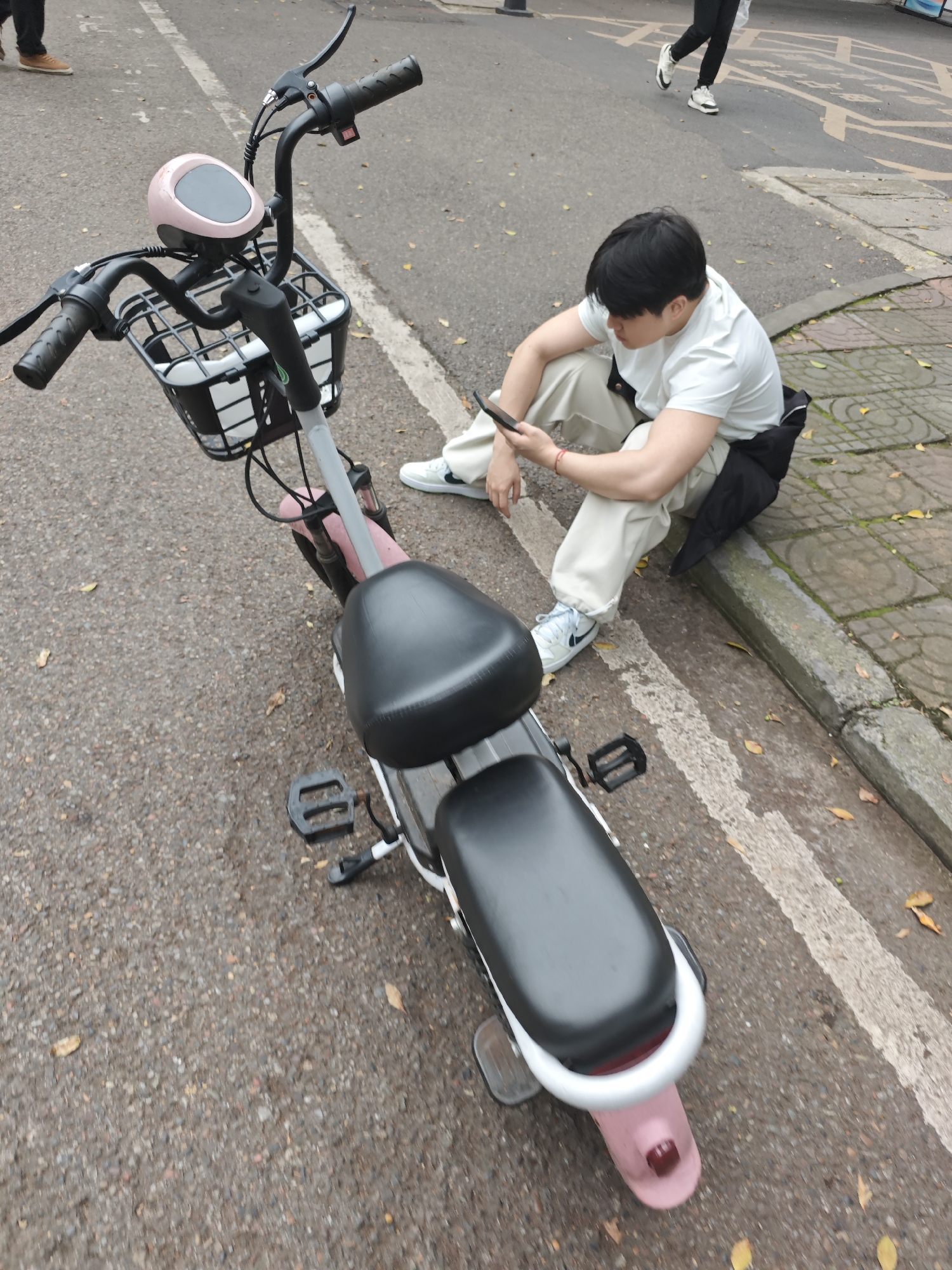 The image shows a person sitting on the edge of a road in what appears to be Chongqing High-tech Zone, China, based on the local surroundings. A pink and white electric bicycle with a basket is parked near the person. The person is looking at a mobile phone. The road is marked with white lines and there are trees and paving stones visible nearby. There is a pedestrian walking in the background.