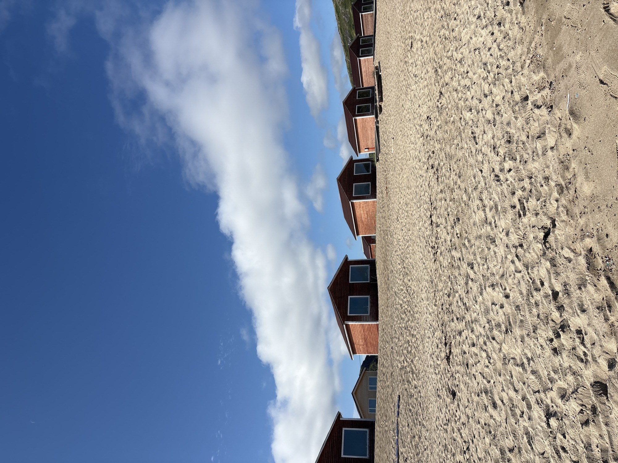 A low-angle, eye-level shot captures a line of small, identical wooden cabins with red siding and white trim situated on a sandy beach. The cabins are positioned in a row, facing the viewer, with visible windows that appear to be reflecting the sky. The beach itself is composed of light brown sand with visible footprints and some scattered debris. Behind the cabins, a gently sloping, treeless hill covered in sparse green vegetation rises towards the horizon. The sky above is a vibrant blue, dotted with fluffy white clouds. The image appears to be taken in Teriberka, Russia, a coastal village known for its remote, natural landscapes.