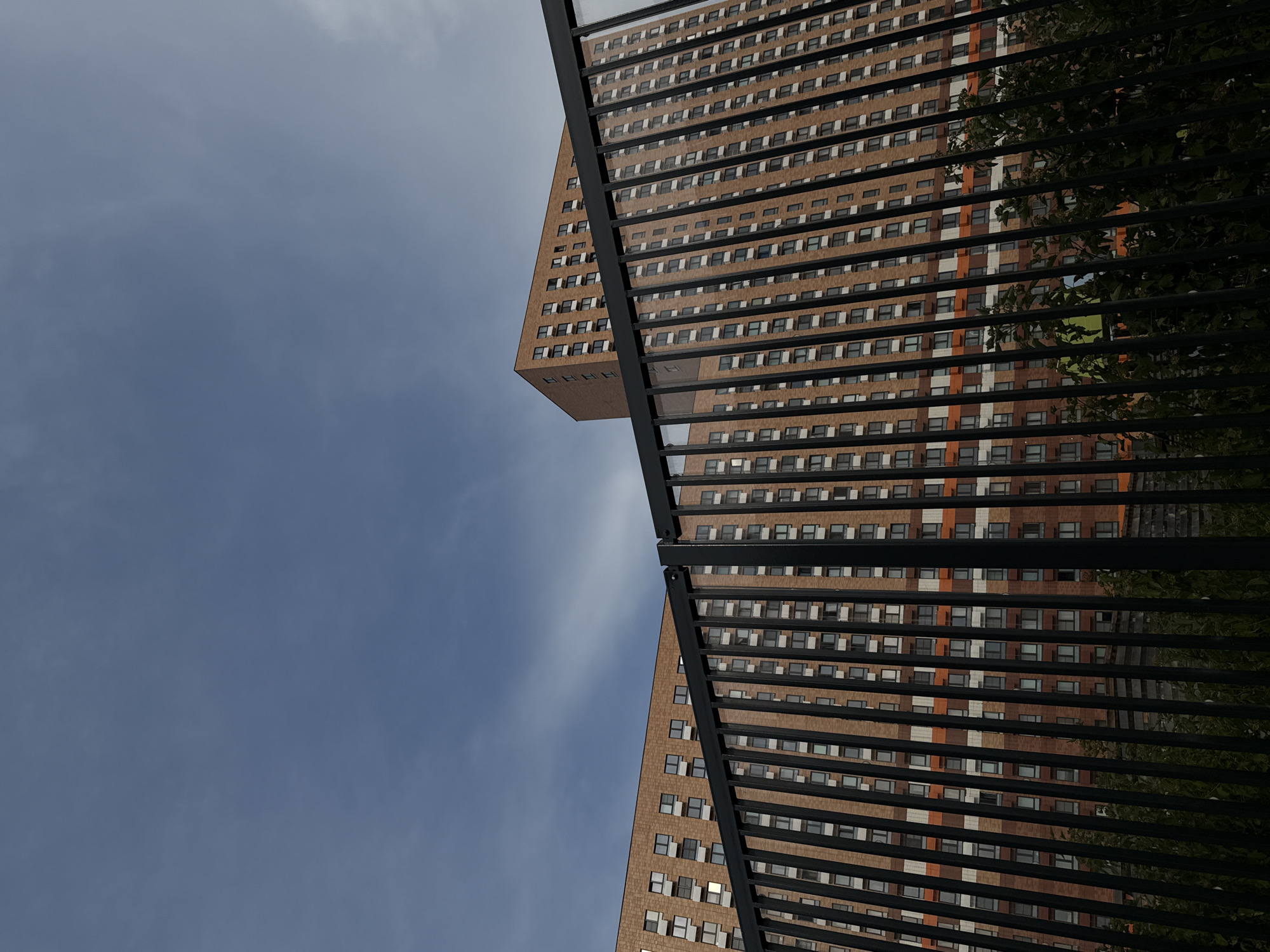 A low-angle shot captures two modern apartment buildings made of light brown brick under a cloudy blue sky. A black metal fence runs across the foreground, obscuring the lower portions of the buildings and some dark green foliage. The buildings feature repetitive patterns of windows with white frames. One building is a long, continuous structure, while the other is a taller, more angular tower with a visible zigzag pattern on its facade. The overall scene suggests an urban residential area in Moscow, Russia.
