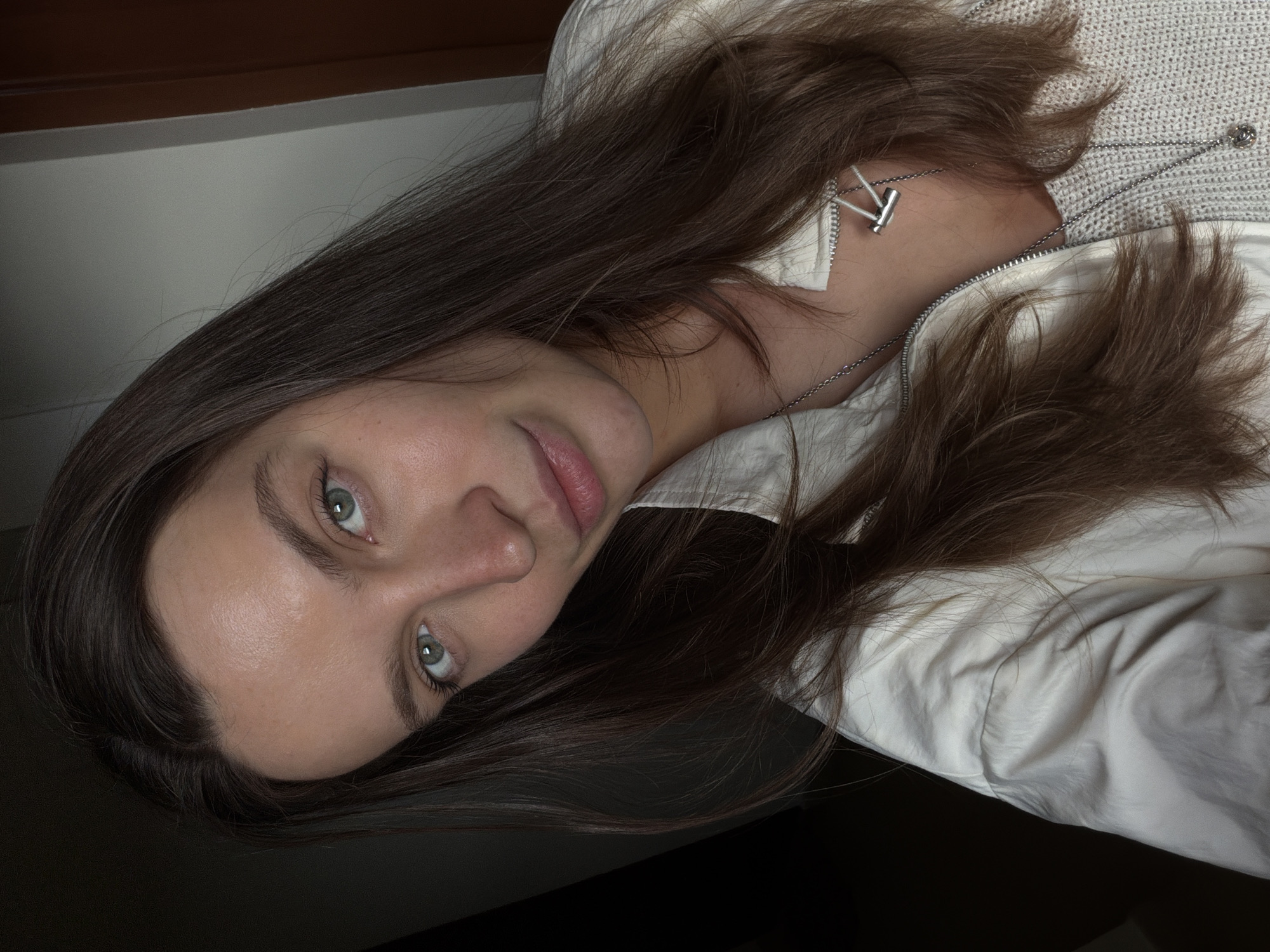 The image, presented at a 90-degree counter-clockwise rotation, features a close-up portrait of a young woman. She has long, dark brown hair that spreads across the frame, highlighting her face. Her skin is fair, and her captivating eyes are a striking shade of green-blue, directed intently towards the viewer. Her expression is calm and serene, with a slight, relaxed smile and naturally pink, slightly parted lips.
She is dressed in a light-colored, possibly white or off-white, collared jacket or shirt, which is partially unzipped. Beneath it, a textured, light-grey knitted garment is visible. A silver chain necklace, adorned with a distinctive T-bar clasp, rests against her neck and the knitted fabric.
The background is dark and indistinct, suggesting an indoor setting with low ambient lighting, possibly in the evening or night. The soft lighting illuminates her face and hair. No specific location identifiers for Budva, Montenegro, are visible within the frame. There is no discernible text in the image.