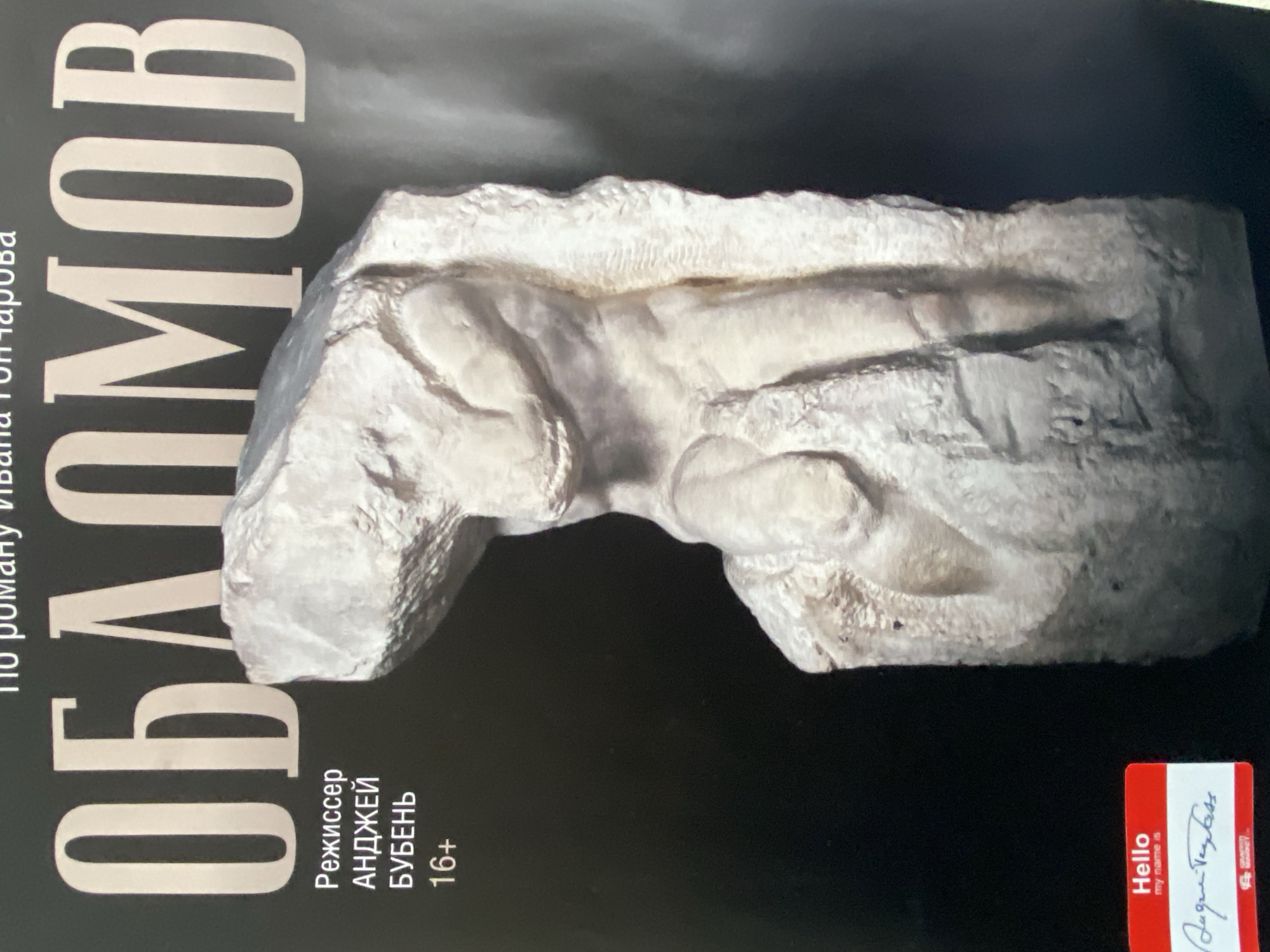 The image shows the cover of a book or poster titled "Oblomov," based on the novel by Ivan Goncharov. The text "Oblomov" is written vertically in large, white, block letters on the left side of the image. There's additional text to the left of that, also written vertically, but it's difficult to read. Beneath the title "Oblomov" is the text "Режиссер АНДЖЕЙ БУБЕНЬ 16+", which translates to "Director Andrzej Buben 16+". In the upper right area of the image, there's a sculpted, white object, possibly a plaster model or a fragment of a sculpture. In the lower-right corner is a red "Hello My name is" name tag. The background is a dark black color. The information about the city of Шебунино, Russia, is not visibly available in the provided image.