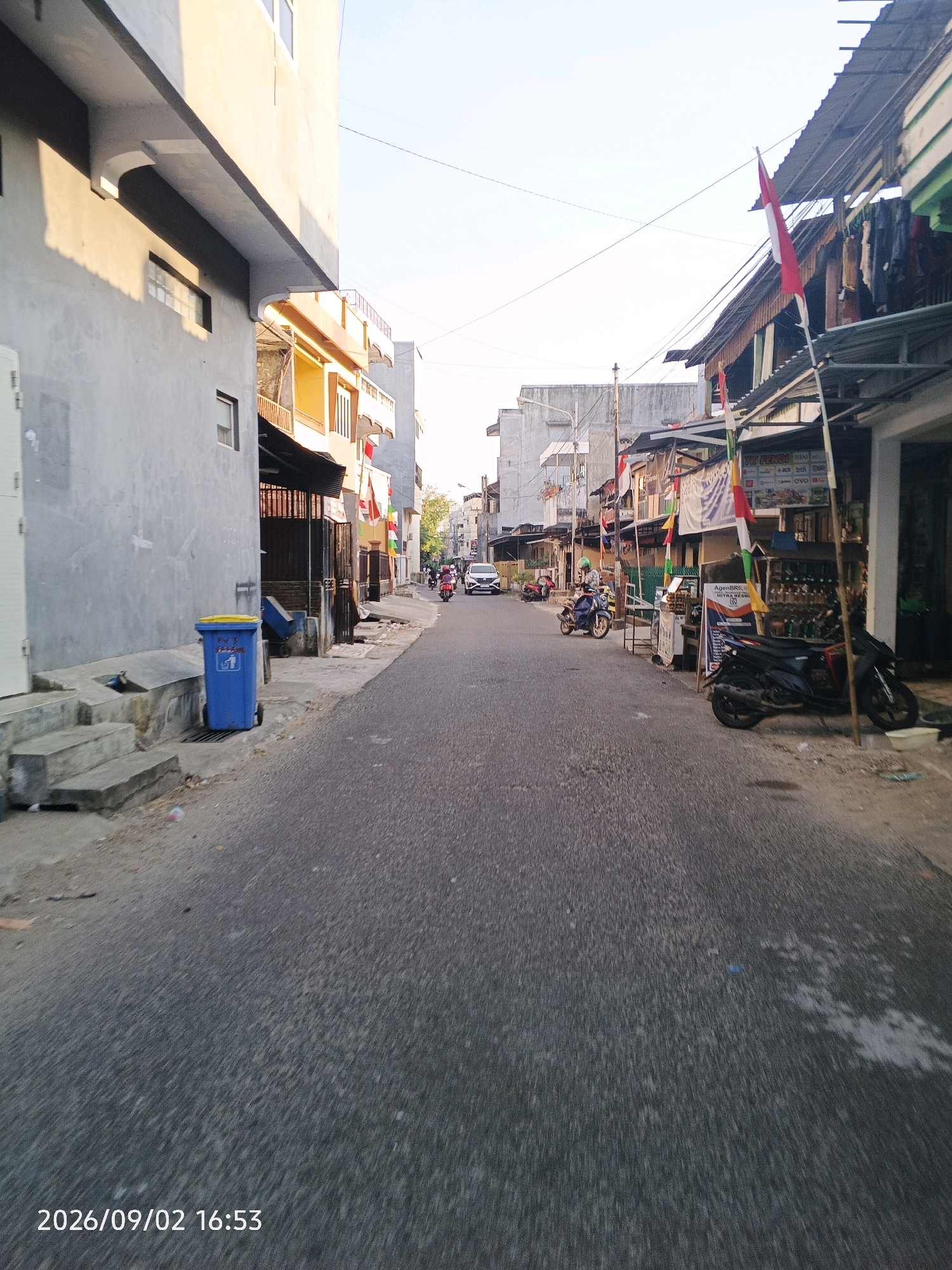 The image depicts a street scene in Pare-Pare, Indonesia, on September 2, 2026, at 16:53. The perspective is from the street, looking down a paved road lined with buildings. On the left, a light gray building with some architectural details and windows is prominent. A blue trash bin with a yellow lid sits on the sidewalk in front of this building. Further down the street on the left, there are more buildings with yellow accents and balconies.
On the right side of the street, there are several businesses with awnings and signs. Indonesian flags and banners are displayed along the storefronts. Several motorcycles are parked along the right side of the road. A dark-colored motorcycle is parked in the foreground, and another black motorcycle is further down. A person is seen riding a motorcycle with a green helmet in the middle ground, heading away from the camera. A white car is also visible in the distance, moving in the same direction. The sky is clear and bright, suggesting it is late afternoon. The overall scene conveys a typical urban street environment in Indonesia, possibly in a commercial or residential area.