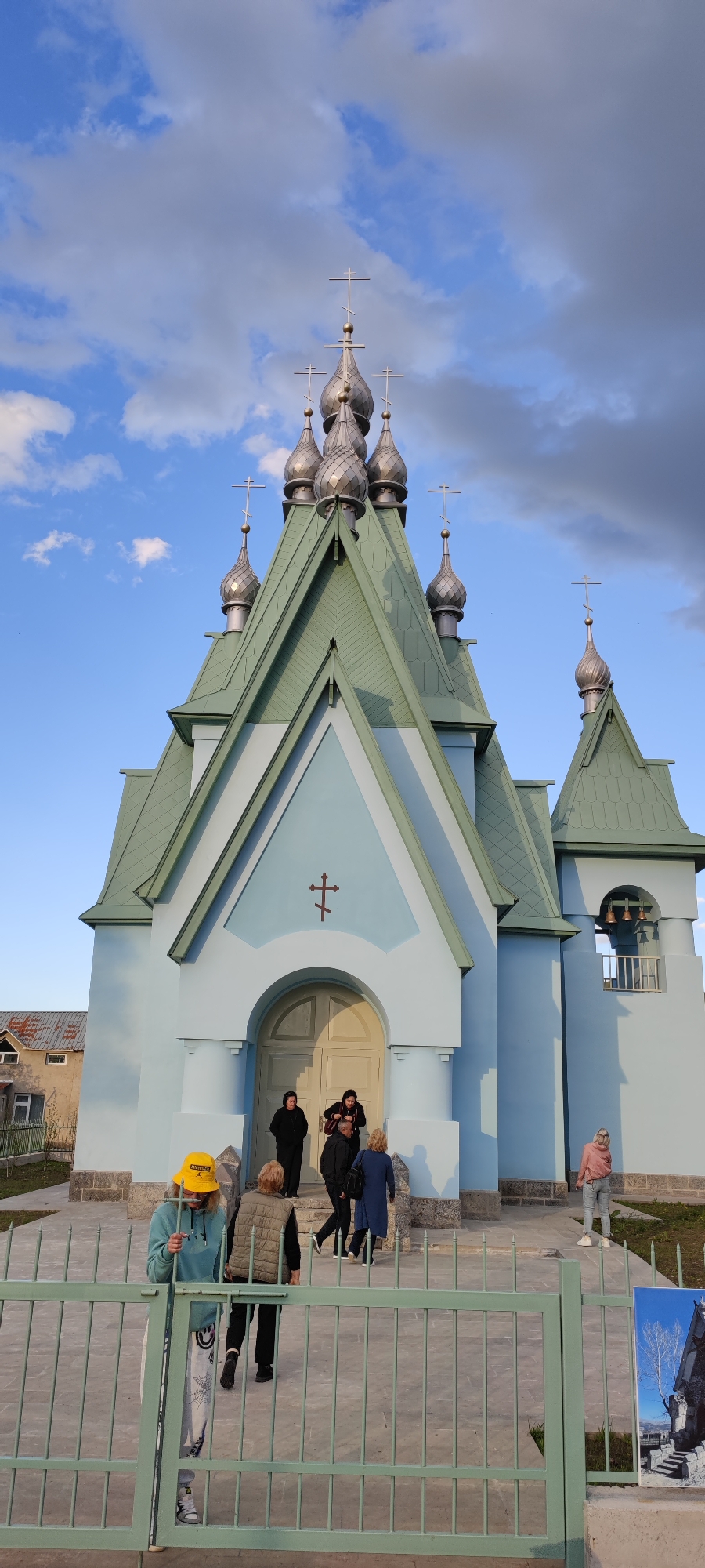 The image captures an Eastern Orthodox church in Amrakits, Armenia, with several individuals present. The church features a light blue facade, green roofing, and distinctive silver onion domes topped with Orthodox crosses. The entrance is framed by an arched doorway with a simple cross above.
People are visible in front of the church. Near the entrance, a group is gathered, including individuals dressed in dark clothing. A person wearing a yellow hat and casual attire stands near a green metal fence in the foreground. Another person is standing near the corner of the building.
The environment includes a partly cloudy sky, with a mix of blue and gray clouds. A building is visible in the background.