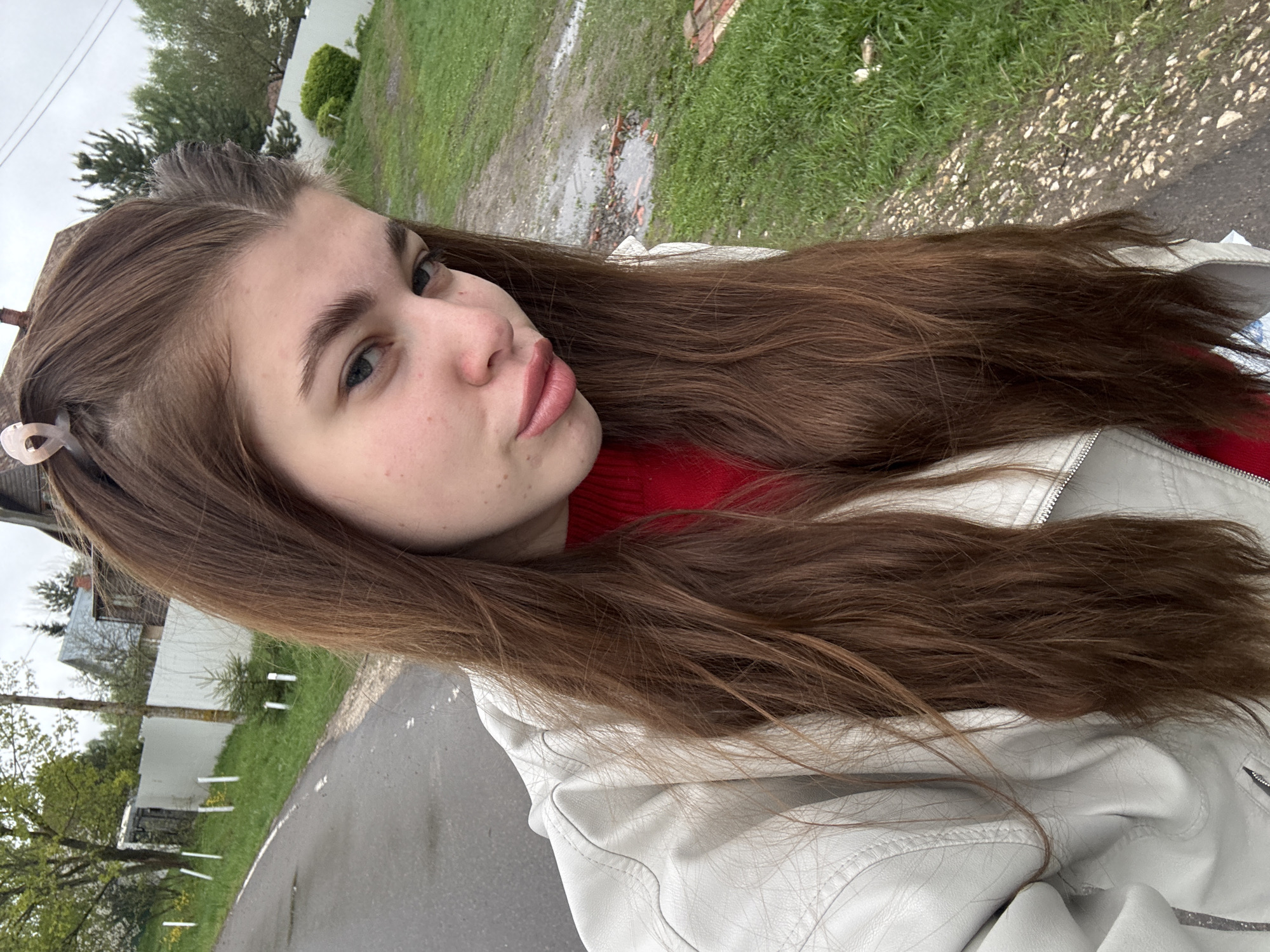 A young woman is taking a selfie outdoors. She has long brown hair and is wearing a red top and a light-colored jacket. A hair clip is visible in her hair. The background suggests an urban or semi-urban environment, with a road and greenery visible. Buildings and possibly other infrastructure elements can be seen in the distance. The scene appears to be in an area near Мещёры, Russia, based on the surrounding environment, including the types of buildings and vegetation.