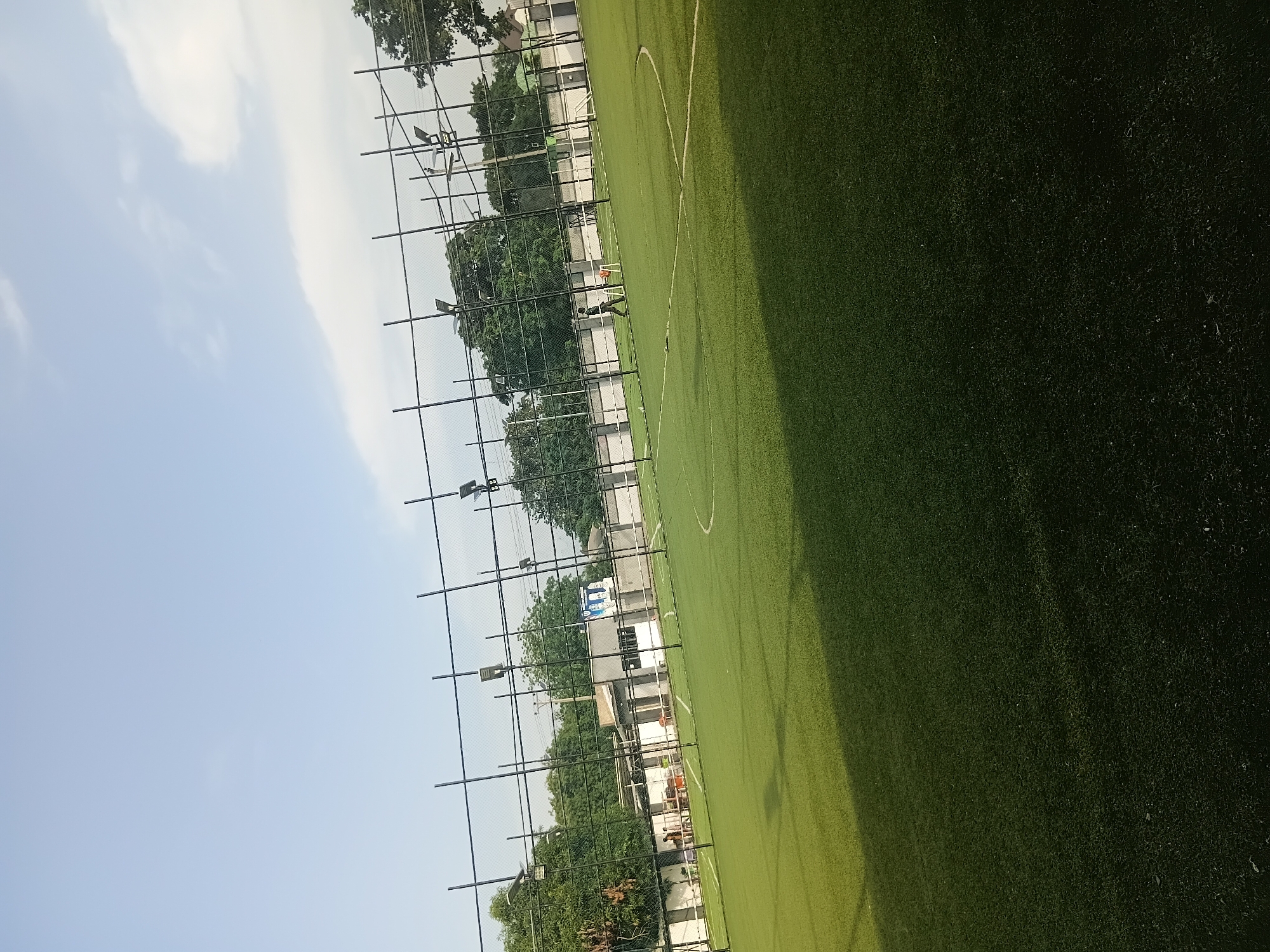 A person is visible on a bright green, artificial turf football field in Jalingo, Nigeria. The field has white markings, including a curved line that appears to be part of a penalty area. A black metal fence surrounds the field, with metal poles extending upwards. Lights are mounted on some of these poles. In the background, there are trees and buildings, suggesting an urban or semi-urban environment. The sky is light blue with some white clouds, indicating it is daytime. The overall scene depicts an outdoor sports facility.