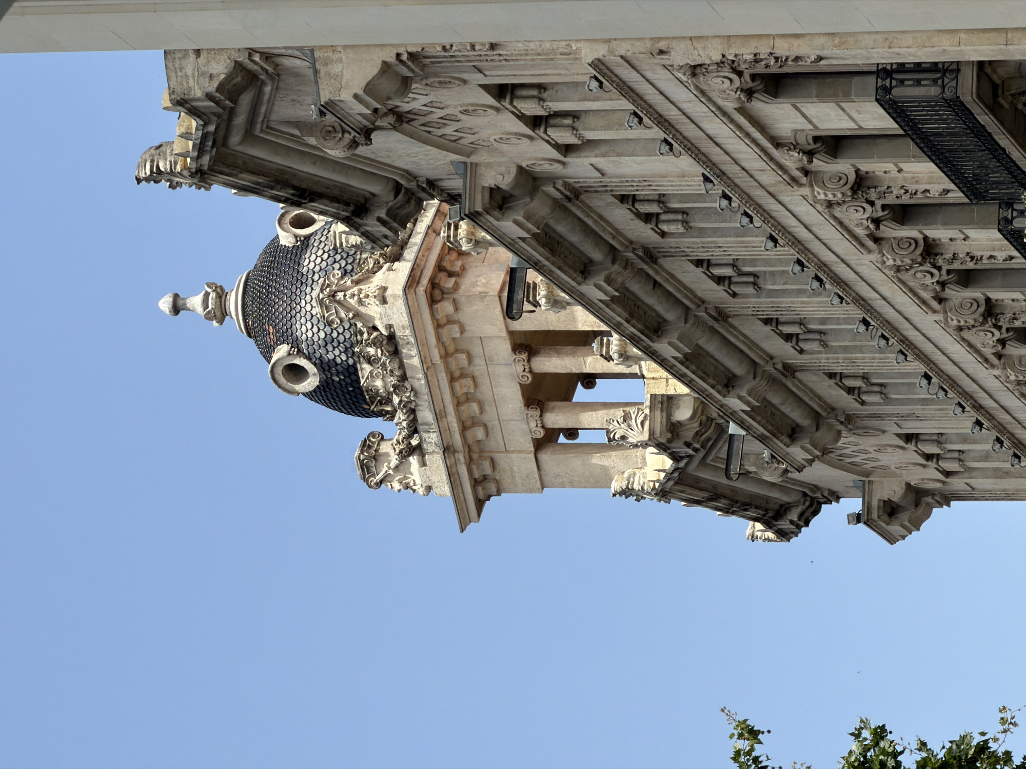 This is a low-angle, rotated photograph of ornate architectural details on a building, set against a clear blue sky. The building's facade features intricate stonework, including decorative moldings, carvings resembling gargoyles, and elaborate cornices. A dark, tiled dome with a finial is prominently visible, adorned with circular elements and sculpted ornamentation. Supporting columns with volute capitals are also present, suggesting a classical architectural style. Lights are attached to the underside of an overhang. In the lower right corner, the tops of green leaves from a tree are visible. The clear sky indicates fair weather, and the lighting suggests it is daytime. The image appears to be taken in a location with significant historical architecture, potentially Barcelona, Spain, given the context provided. No people or specific activities are depicted.