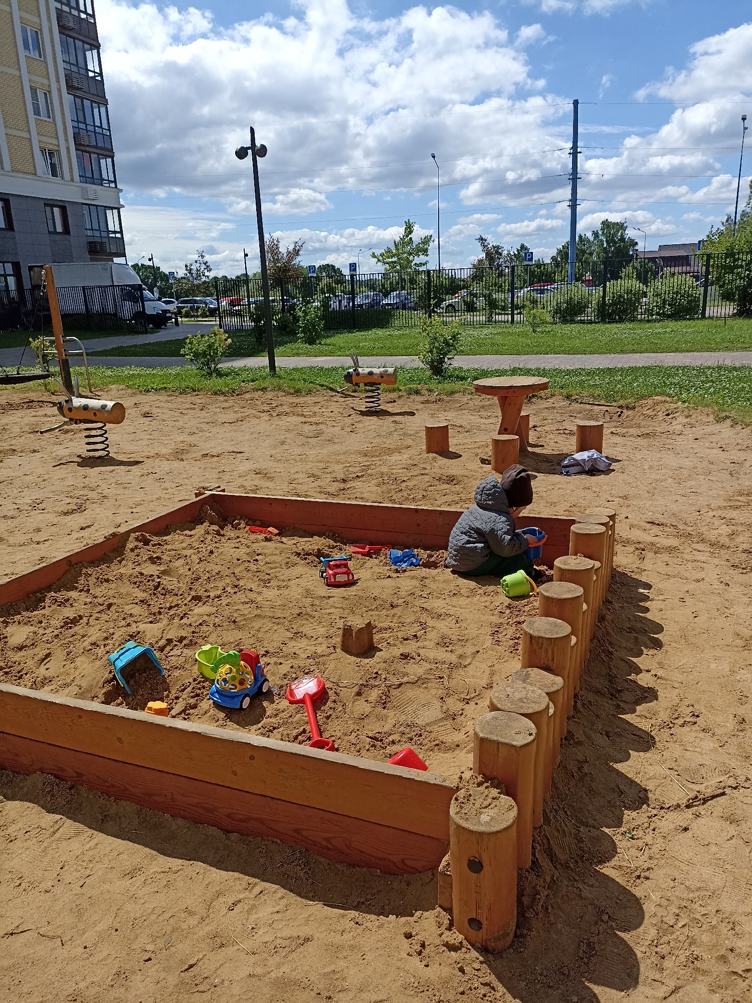 The image depicts a children's sandbox area in the city of Ostafievo, Russia. A young child, wearing a grey jacket and a brown hat, is seated inside the sandbox, actively playing with a blue bucket and sand. Scattered within the sandbox are various colorful plastic toys, including a red toy truck, a green toy with a yellow ball, and a red shovel. The sandbox itself is constructed with wooden planks and is bordered by a series of short, round wooden posts. Outside the sandbox, further into the sandy area, a wooden table with attached stools is visible. The background features a grassy area, a black metal fence, parked cars, trees, and a tall residential building with balconies. The sky is partly cloudy.