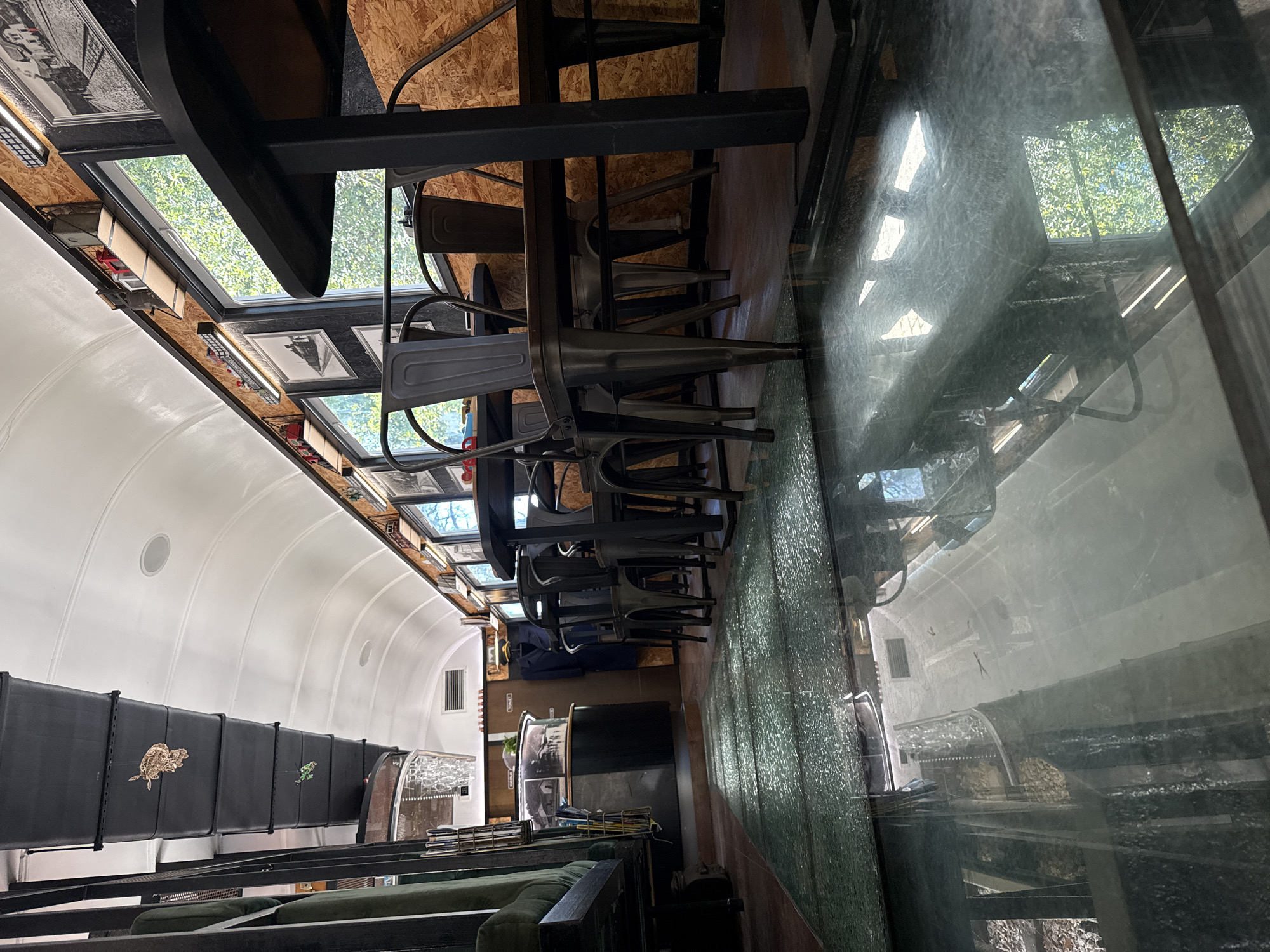 The image shows the interior of a vehicle, possibly a renovated train car or bus, in Sasovići, Montenegro. The space is divided into a dining/seating area and a separate space. The dining area features a glass-topped table with stacked metal chairs. The space above features a series of framed pictures and windows along the top of the car. The seating area is separated by a black partition with decorative brass emblems. A green sofa is situated in this area. A sign indicates the location of a bathroom. A reflective surface on the right side of the image appears to show a distorted view of the opposite side of the vehicle.