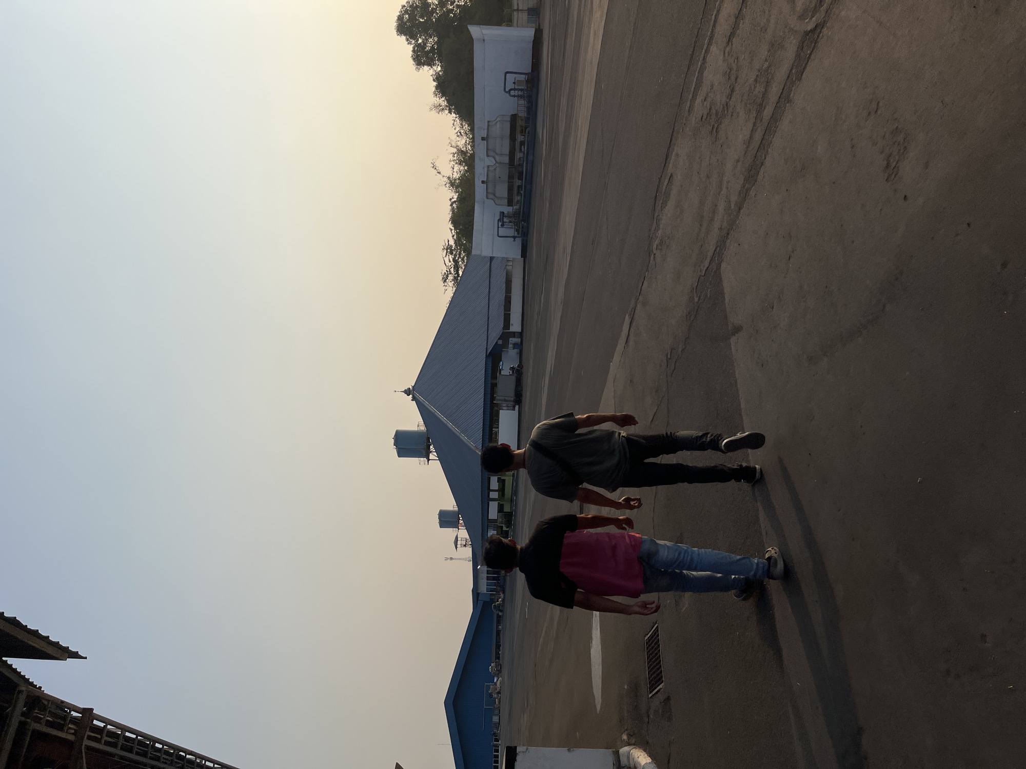 Two individuals are walking away from the camera on an asphalt surface in what appears to be an industrial or logistical area. The person on the left is wearing a pink and black shirt and blue jeans, while the person on the right is wearing a dark gray t-shirt and dark pants. In the background, there are several large, blue-roofed buildings with a distinct, pitched central roof structure, likely a warehouse or factory. Two water towers are visible, one atop a building and another on a separate platform. To the far right, a white structure houses industrial equipment that resembles cooling towers. The sky is clear with a soft gradient of light blue to pale yellow, suggesting either early morning or late afternoon. The environment seems to be an open space with paved ground and some vegetation visible in the distance. The location is identified as Pelang, Indonesia.