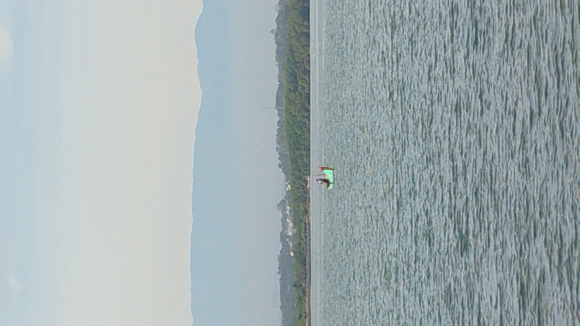 A small, green and white boat with two individuals on board sails on a body of water. The boat has a flag or pole at its rear. The individuals appear to be standing or fishing. The water is calm with small ripples. In the background, a dense line of green trees borders the water. Further back, a hazy, light blue mountain range stretches across the horizon under a pale sky with a few faint clouds. The scene is likely located in the vicinity of Lhokseumawe, Indonesia, given the presence of coastal vegetation and mountainous terrain common to the region.