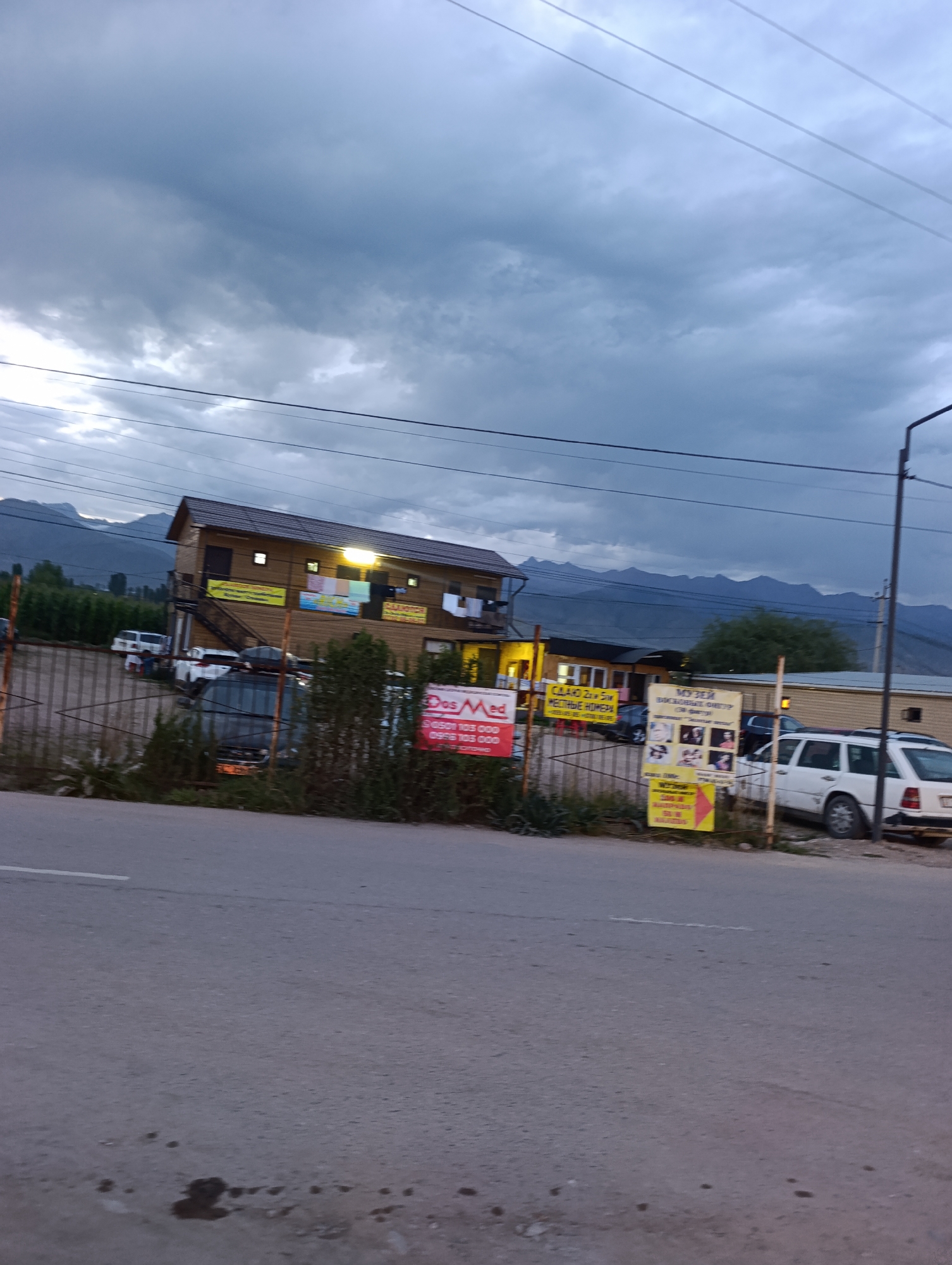 The image depicts a street scene in Bosteri, Kyrgyzstan, under a cloudy sky with distant mountains. A two-story wooden building with illuminated windows stands on the left, a gate with a fence in front of it. Several cars are parked near the building, including a white van, a dark SUV, and a white sedan. Signs are visible on the fence, one red and white with Russian text, and another yellow with Russian text and images. Green trees and vegetation line the area behind the fence. The foreground shows a paved road with some dark spots.